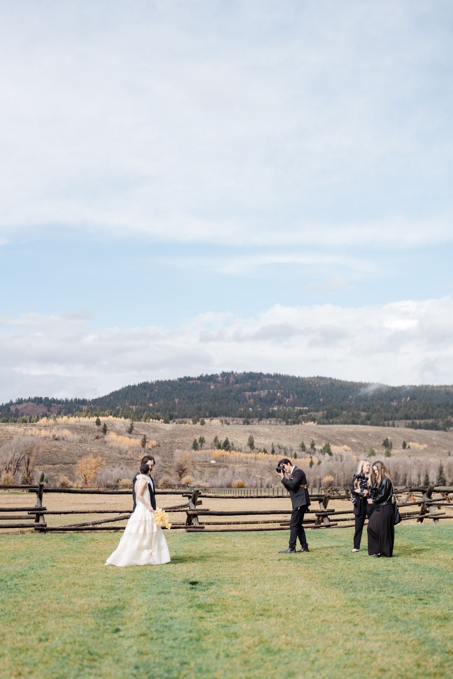 Bride in white gown stands on green field with wedding party near rustic fence, mountains and autumn foliage in background.