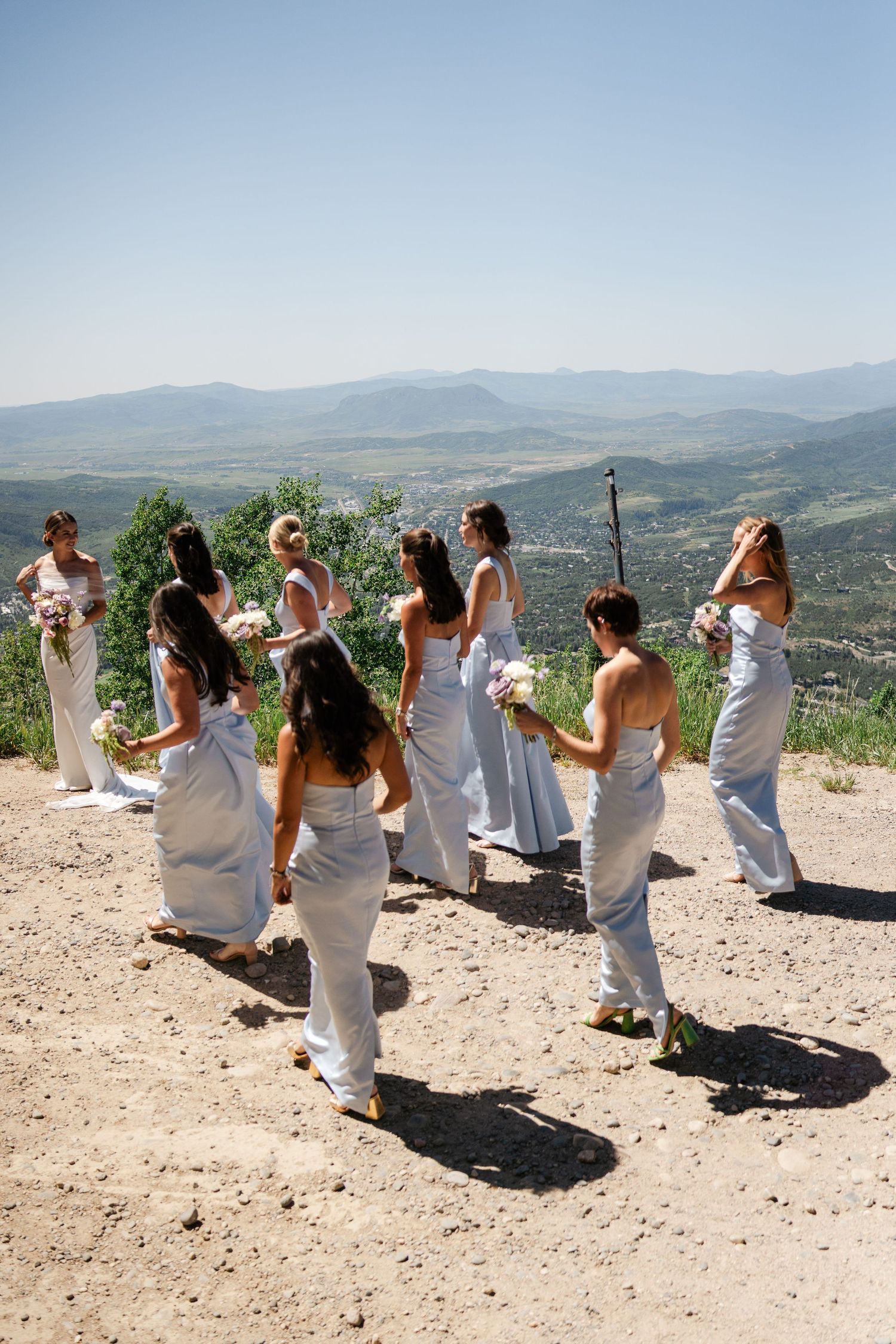 Bridesmaids in light blue gowns walking along a hilltop with sweeping mountain valley views.