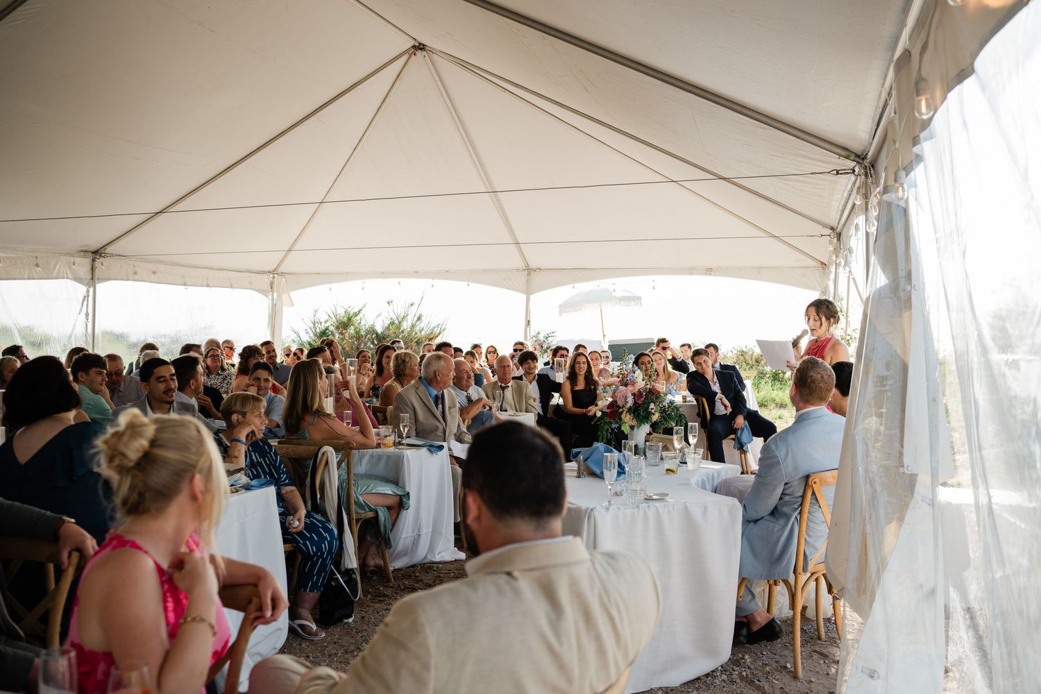 Wedding guests seated at tables listen to a speech during an outdoor tented reception.