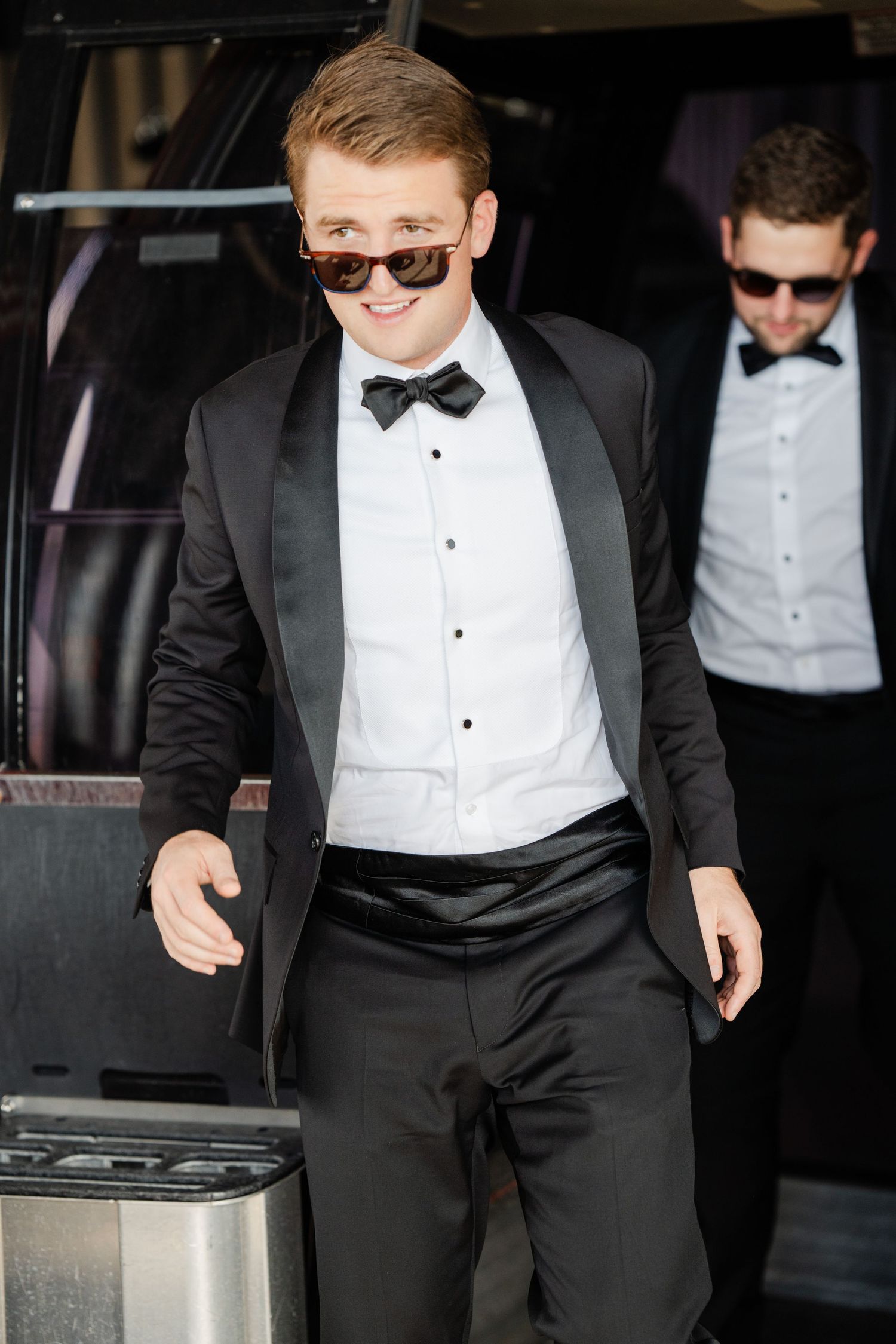 Stylish groom in black tuxedo and sunglasses stepping out of a limousine on wedding day.