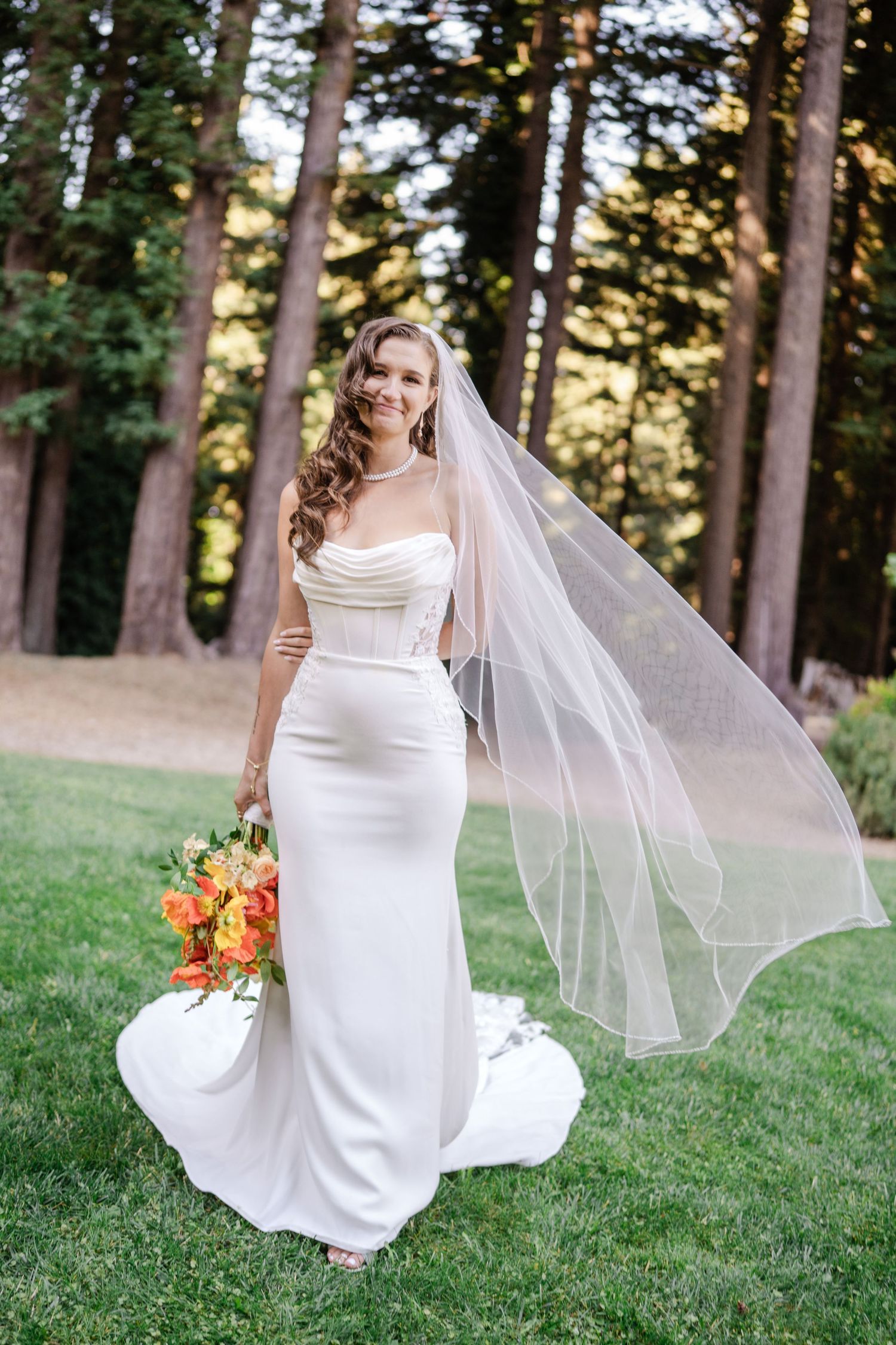 Bride in white gown with flowing veil holding colorful bouquet among tall trees.