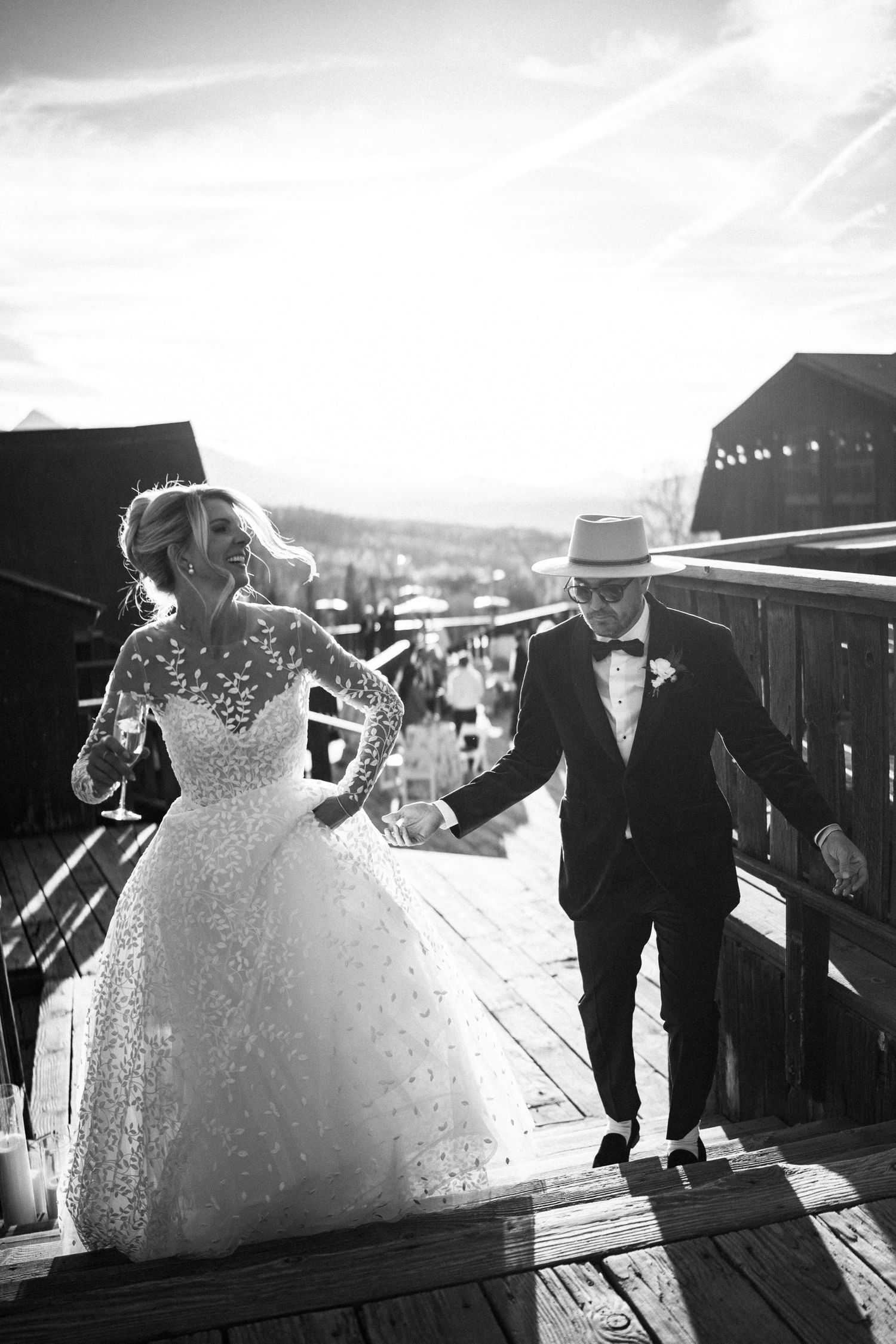 Black and white photo of laughing bride in lace gown and groom in hat on wooden deck.