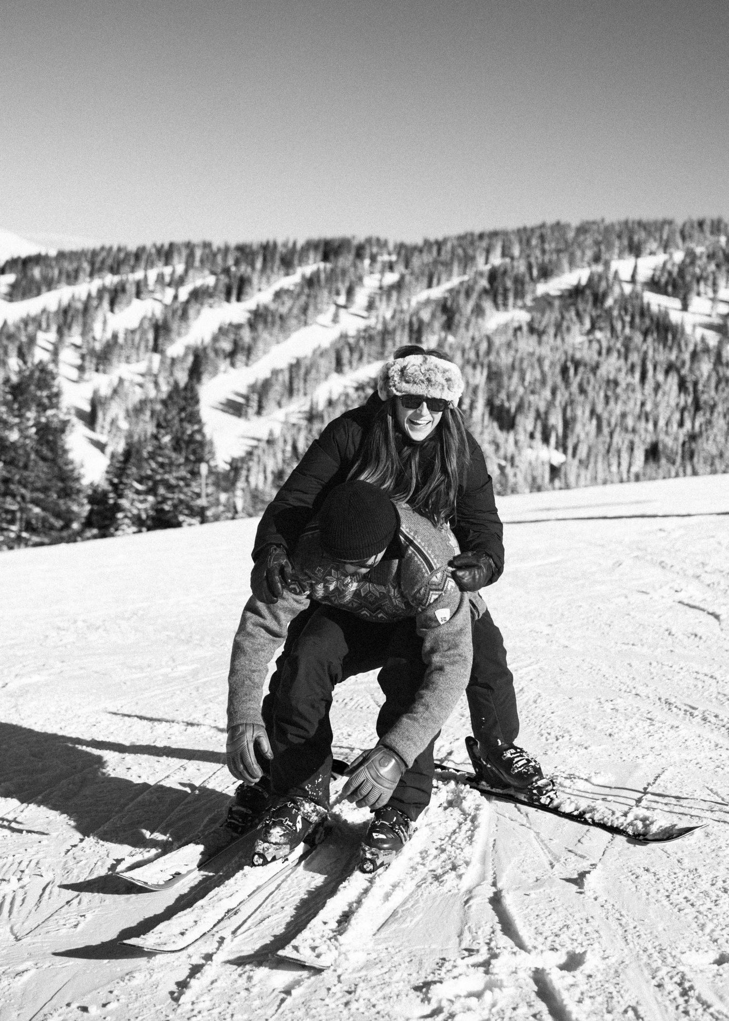 Black and white photo of smiling skier crouching on slope with mountain runs behind.