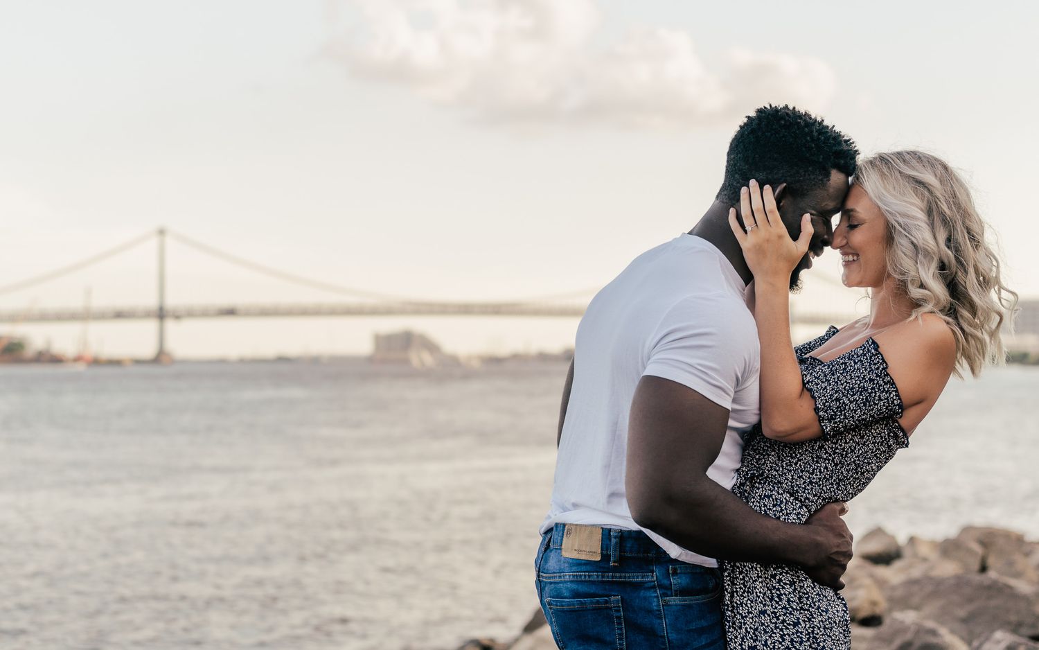 An interracial couple shares a romantic almost-kiss near a waterfront with a suspension bridge at sunset.