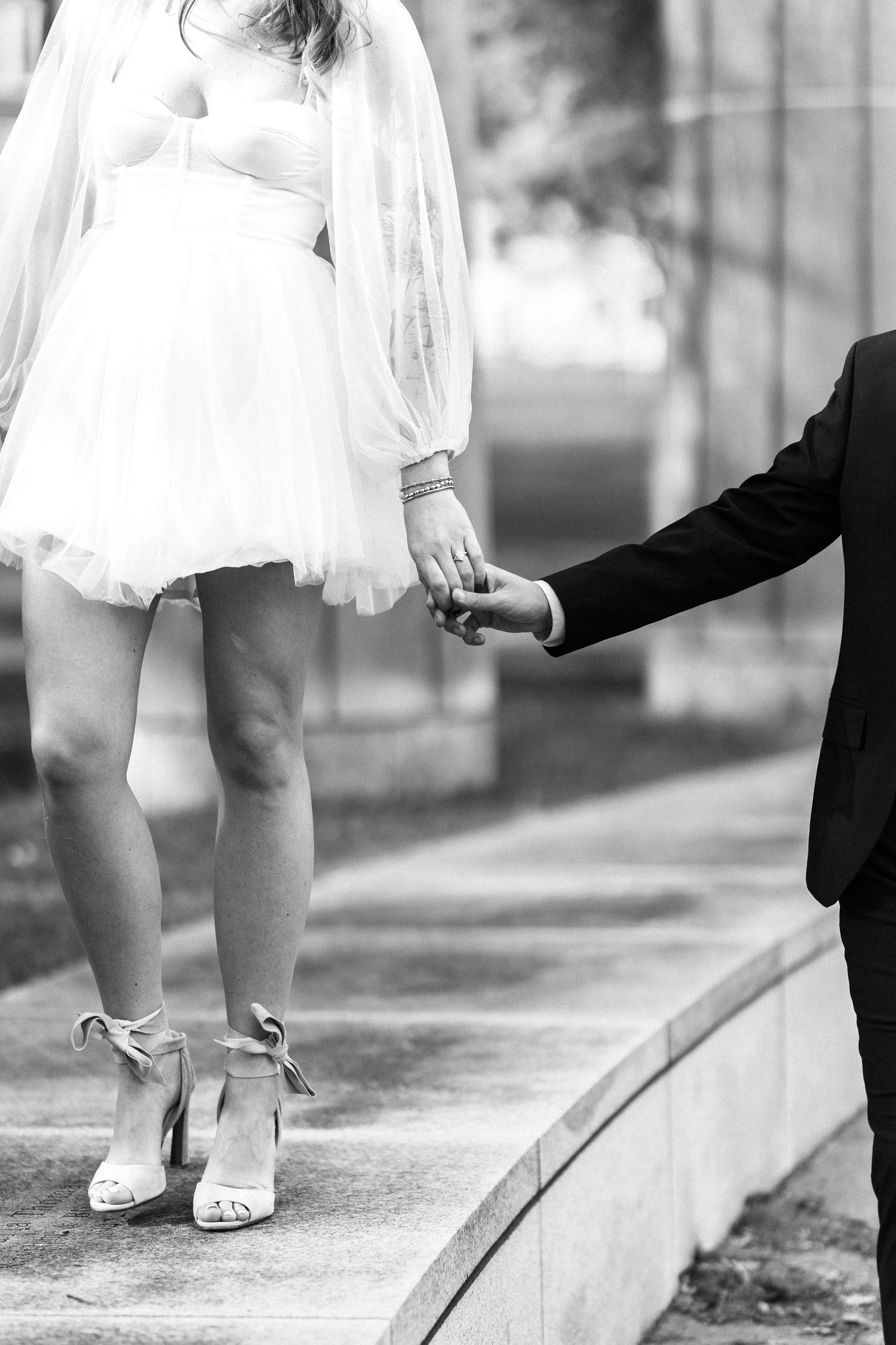 Black and white photo of bride in short white dress holding grooms hand while standing on steps.