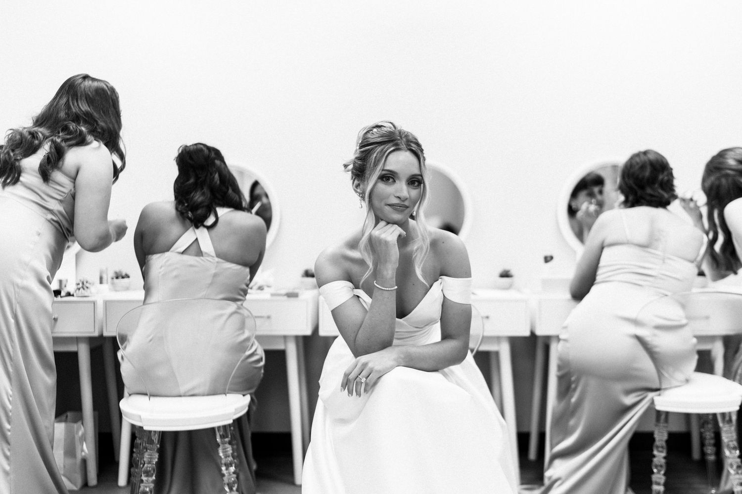 Black and white photo of bride sitting pensively while bridesmaids chat at getting ready table.