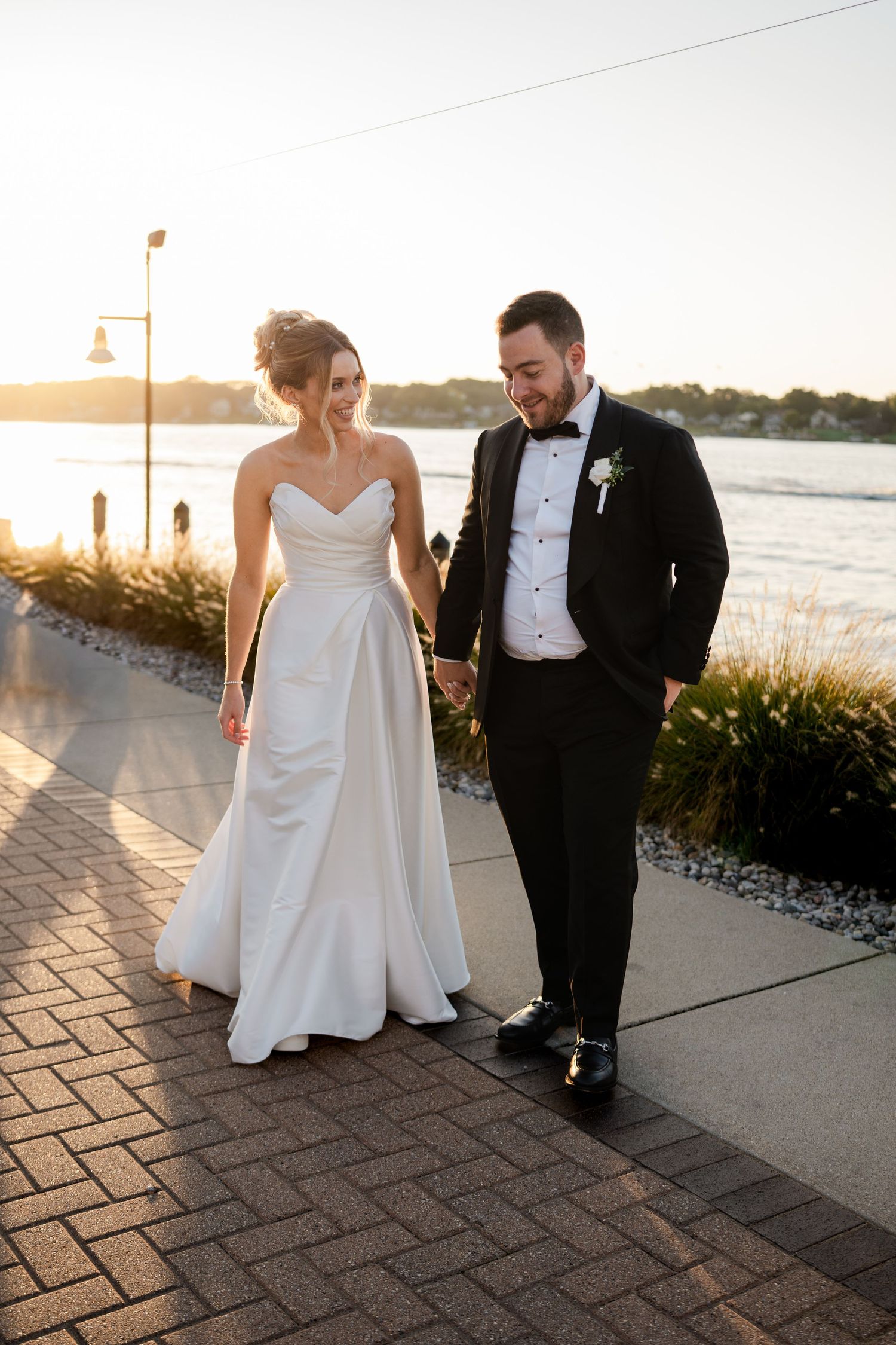 Bride in strapless white gown and groom in tuxedo walk hand in hand along a waterfront path at sunset.