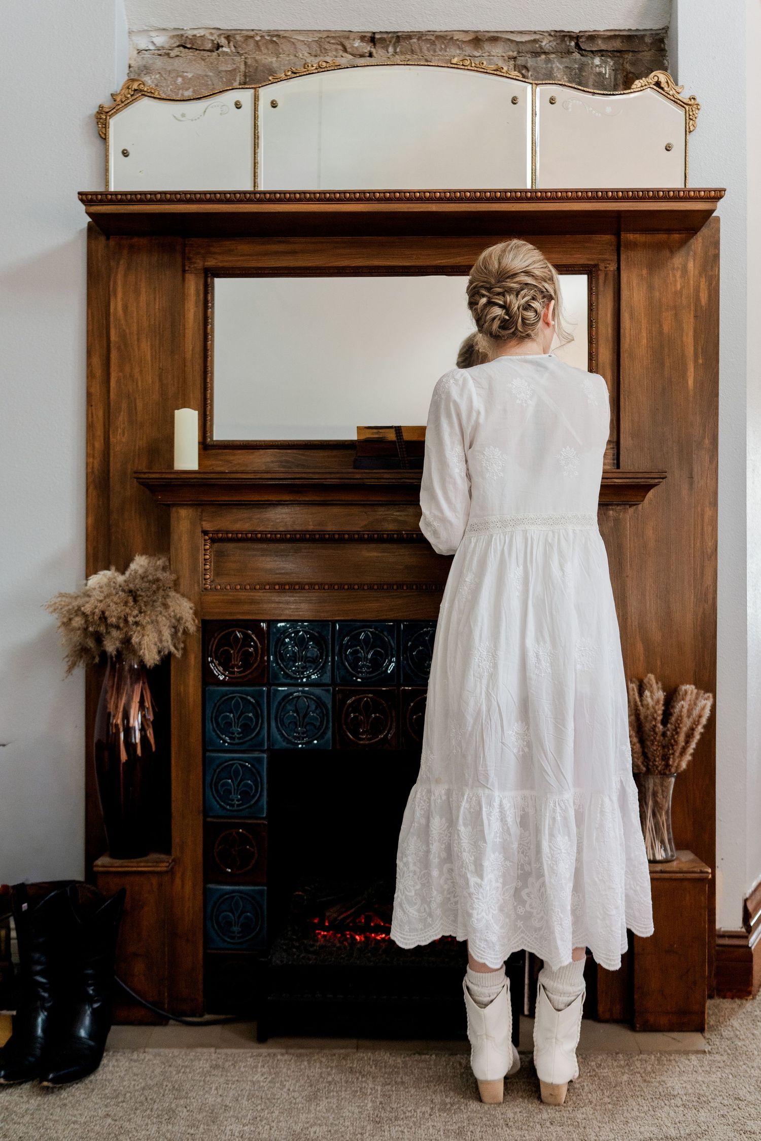 Bride in white midi dress and cowboy boots stands before ornate wooden fireplace with vintage mirror.