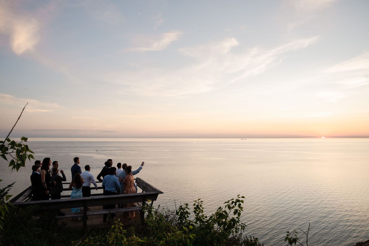 Wedding guests gather on a waterfront deck to watch a stunning sunset over calm waters.