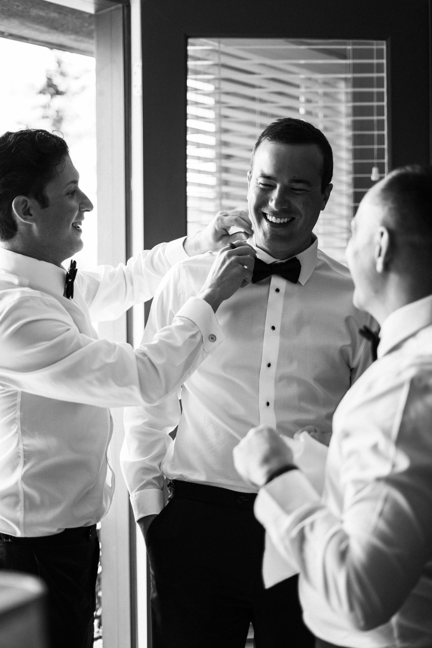 Groomsmen helping groom adjust bow tie while laughing together in black and white wedding preparation photo.