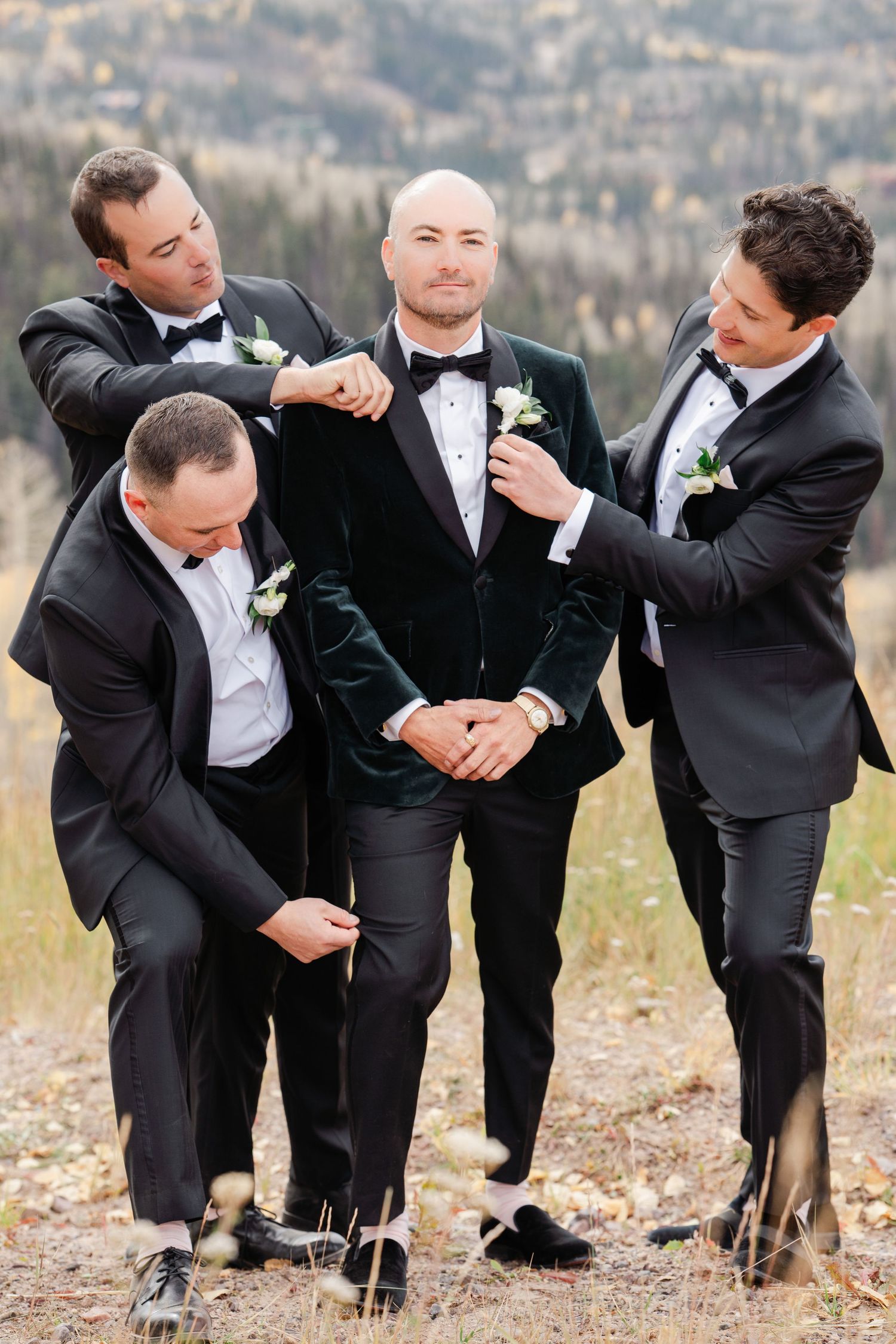 Groom in green velvet tuxedo being helped by three groomsmen in black tuxedos with mountain backdrop.
