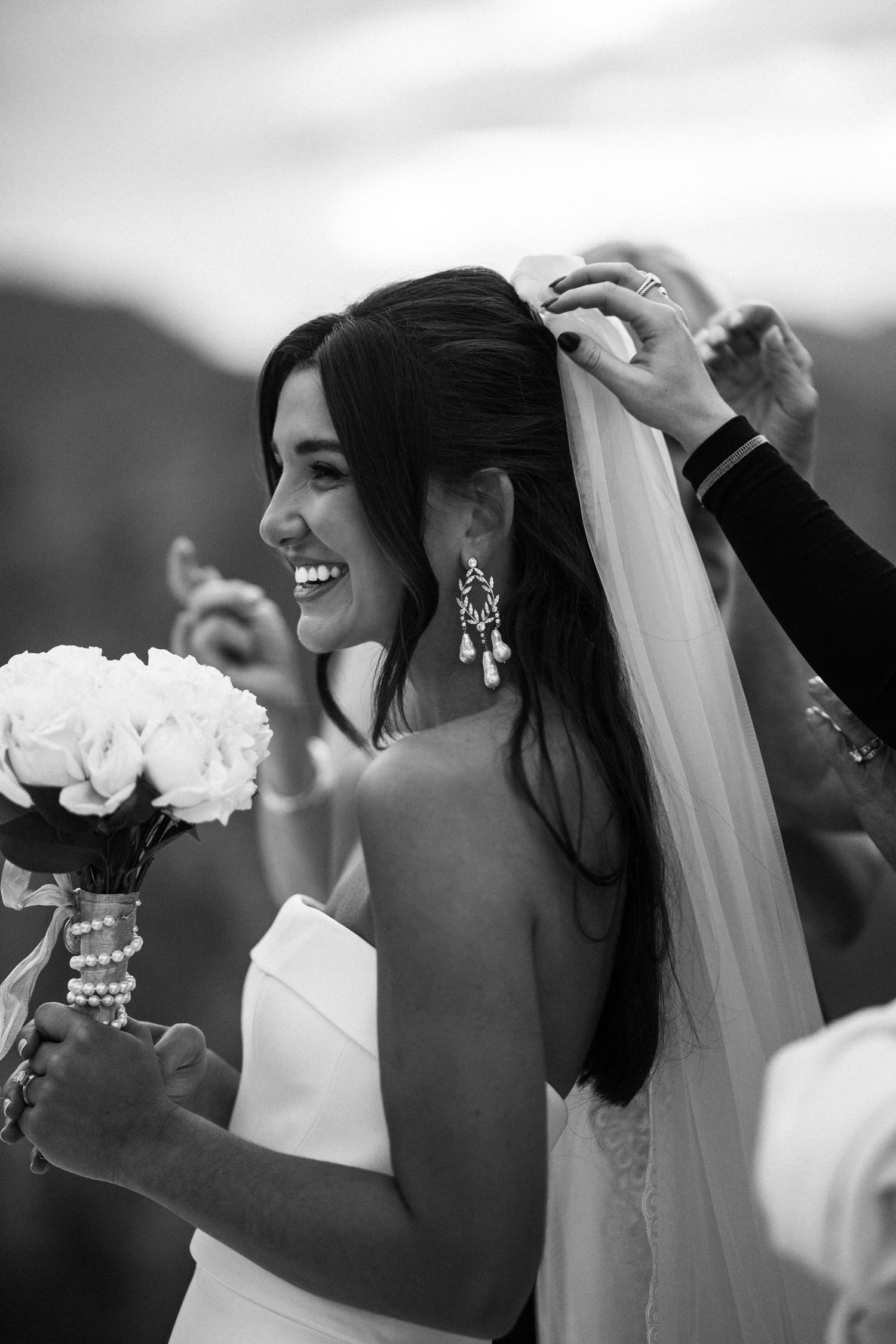 A radiant bride in a white gown laughs while her veil is adjusted at an outdoor wedding ceremony.