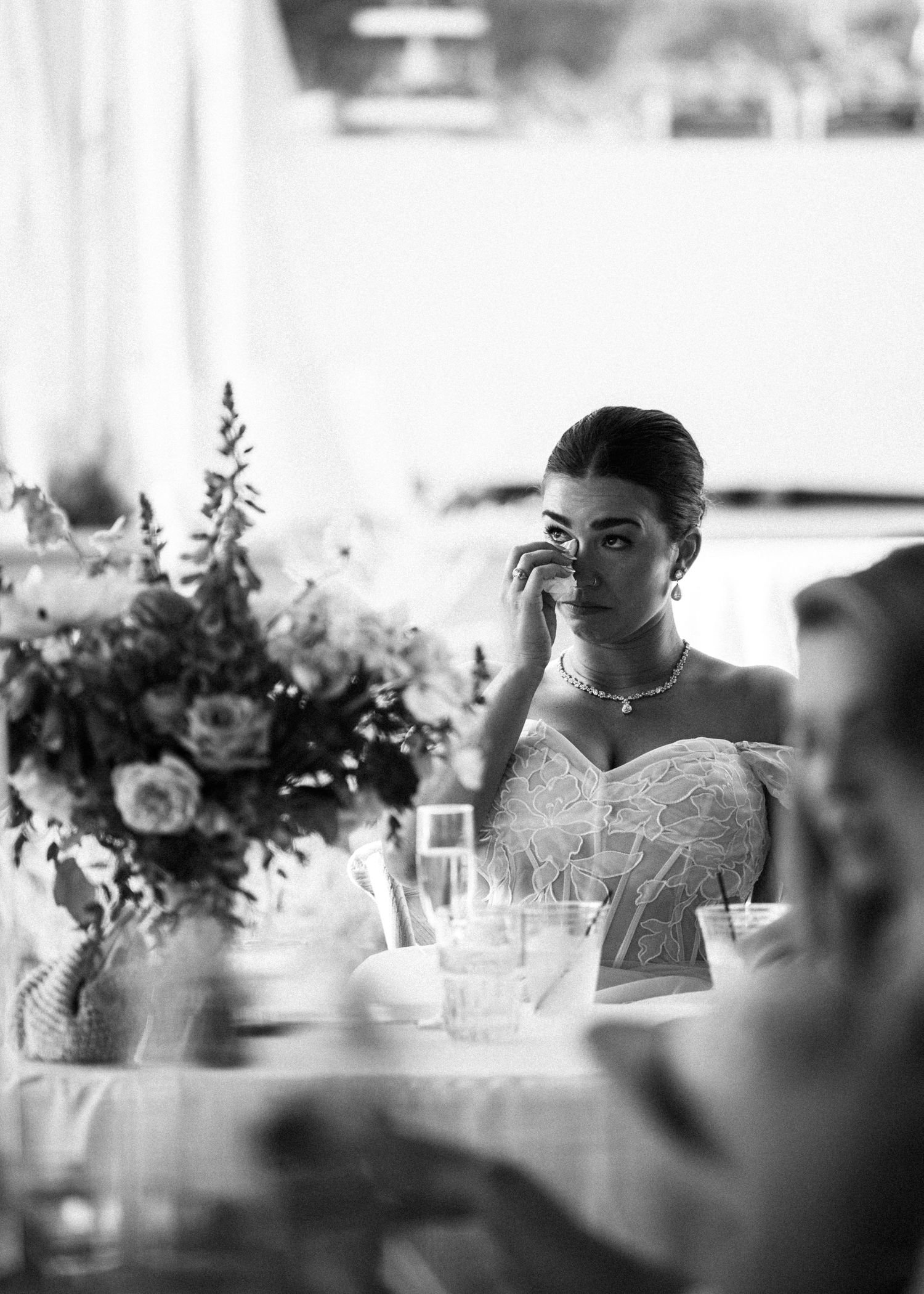 Black and white photo of emotional bride wiping tears at wedding reception table with flowers.