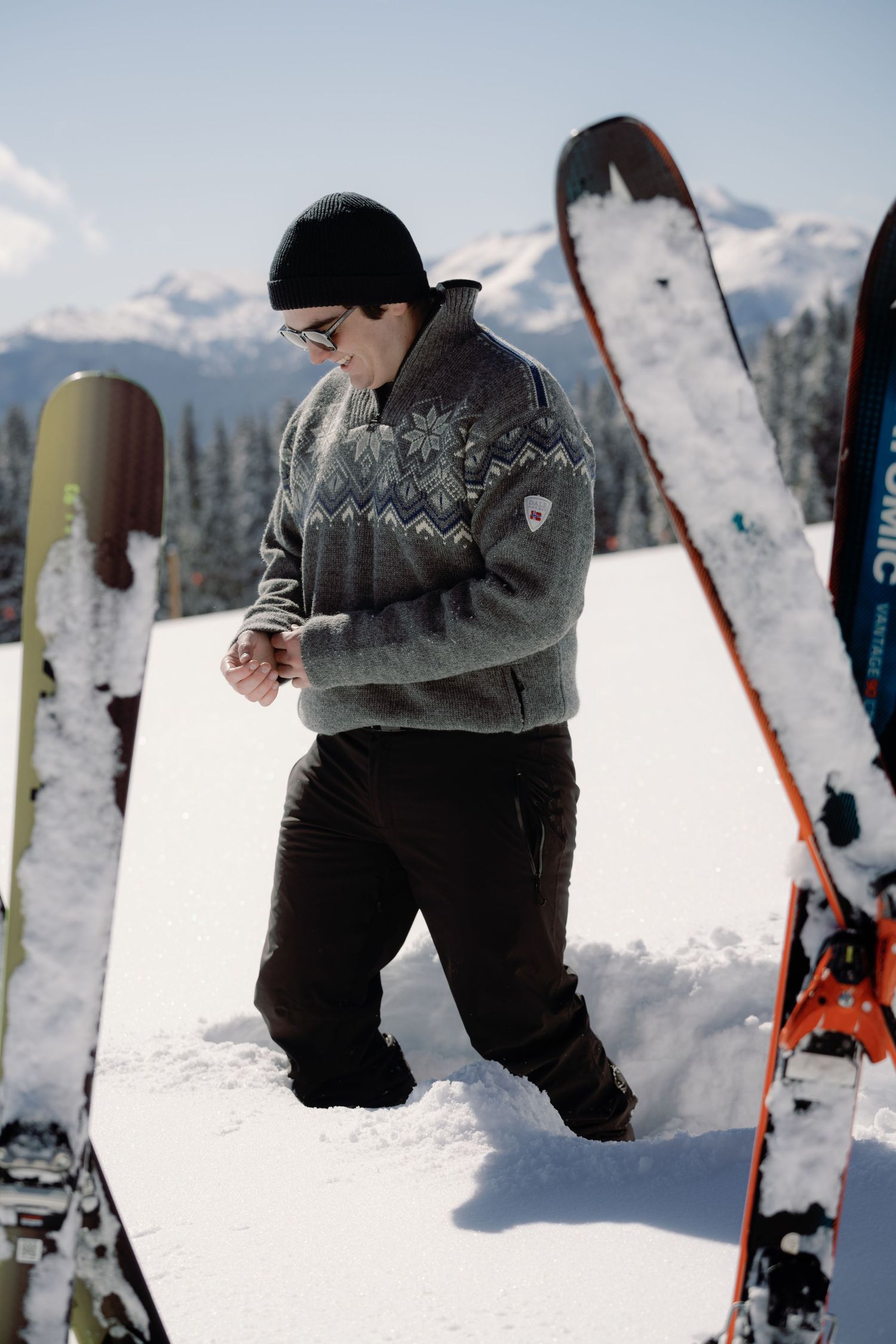 Man in Nordic sweater and sunglasses walking through deep snow beside skis on sunny mountain.