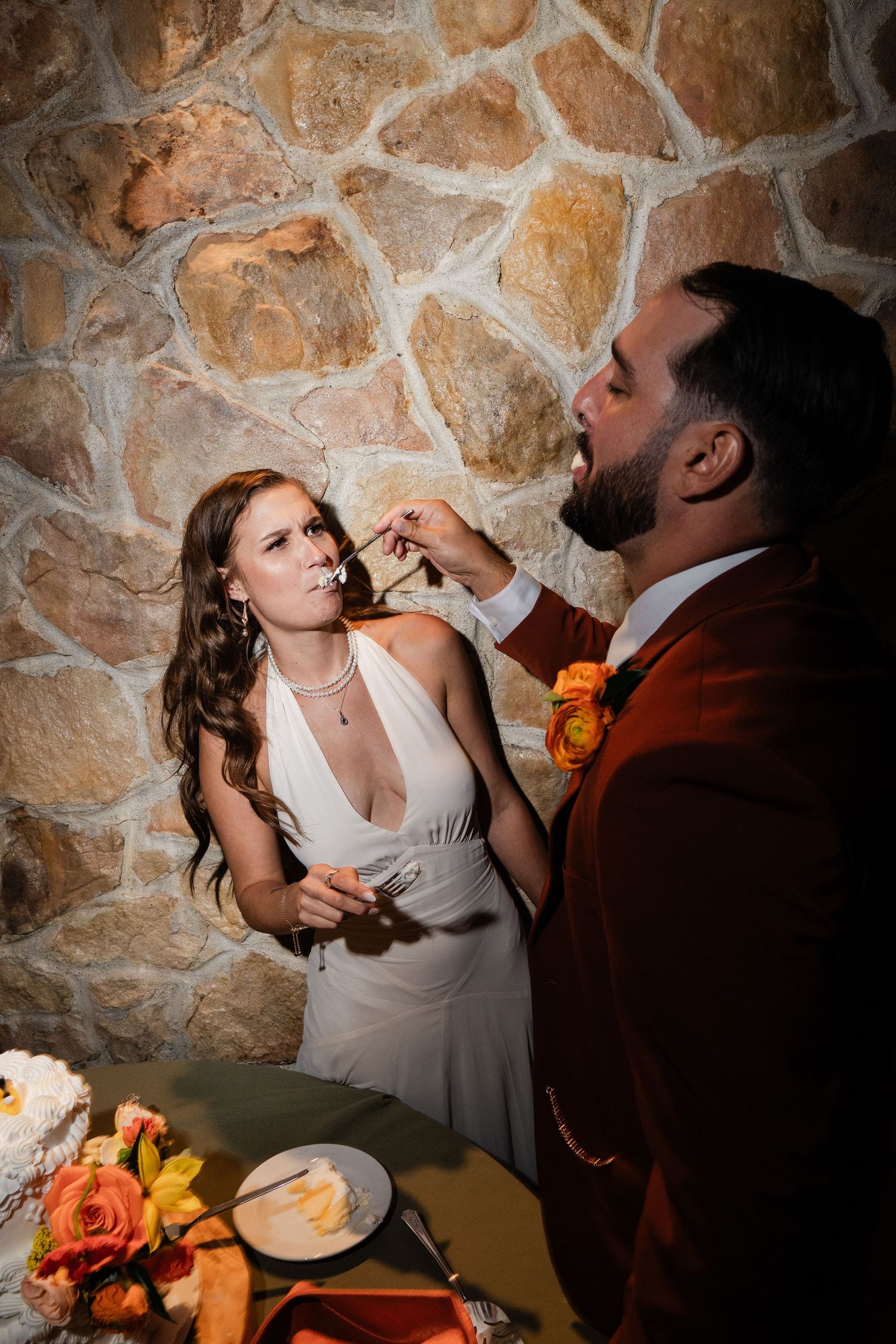 Bride feeds groom wedding cake slice in front of rustic stone wall at reception.