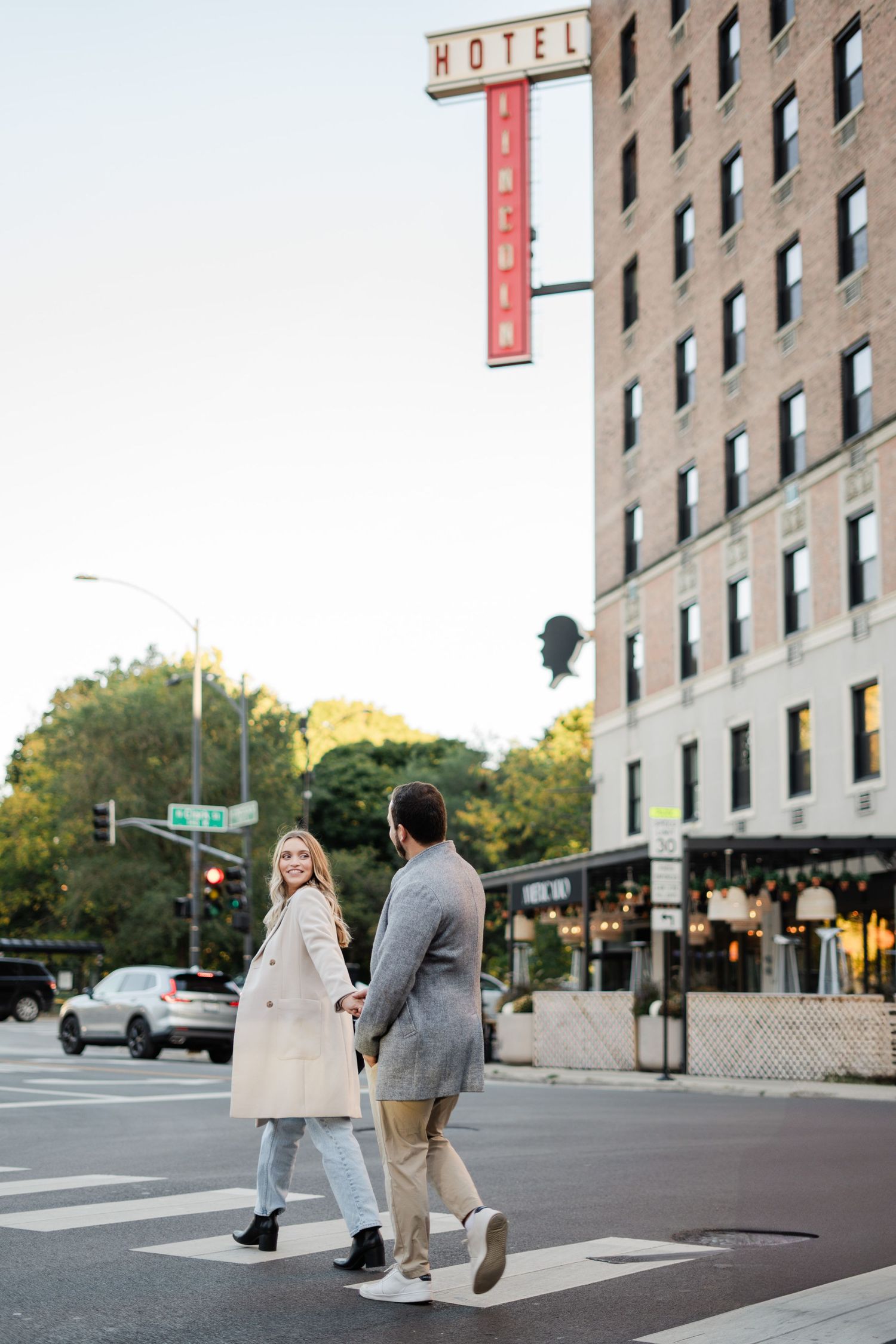 Engaged couple holding hands while crossing a street in front of Hotel Lincoln in Chicago.