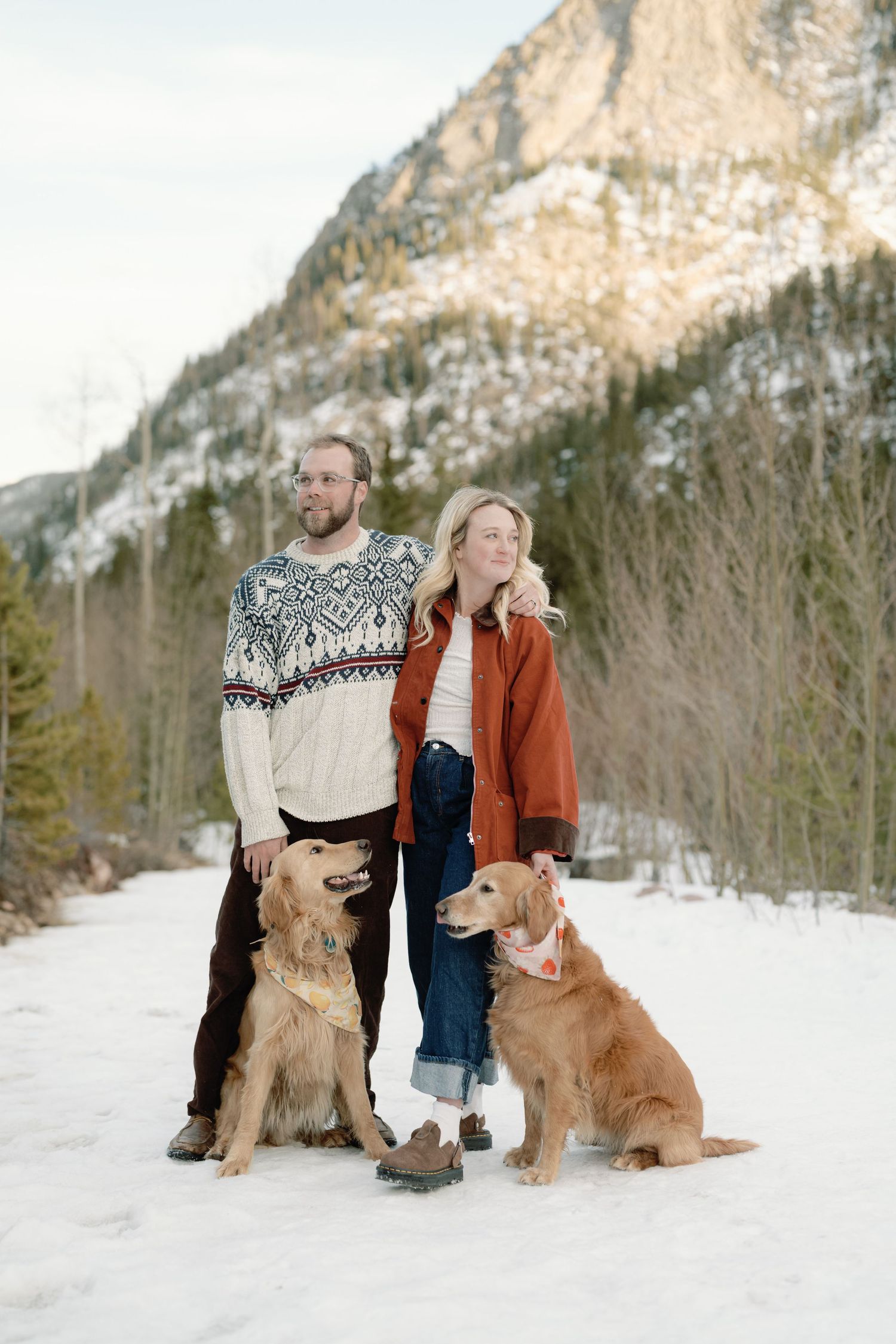 Couple posing with two golden retrievers in a snowy mountain landscape in winter.