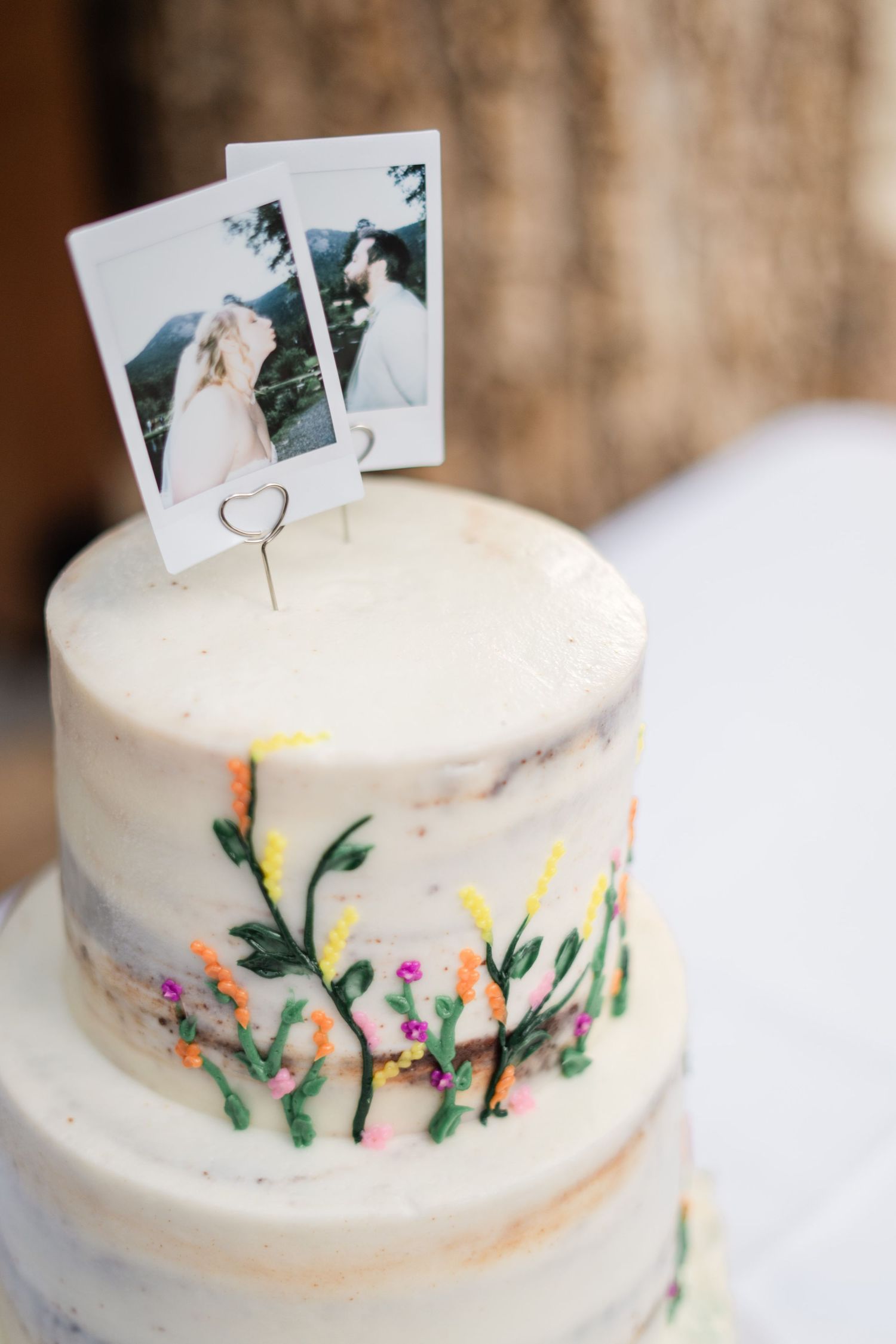 Two-tier white wedding cake with colorful floral details and Polaroid photo cake toppers.
