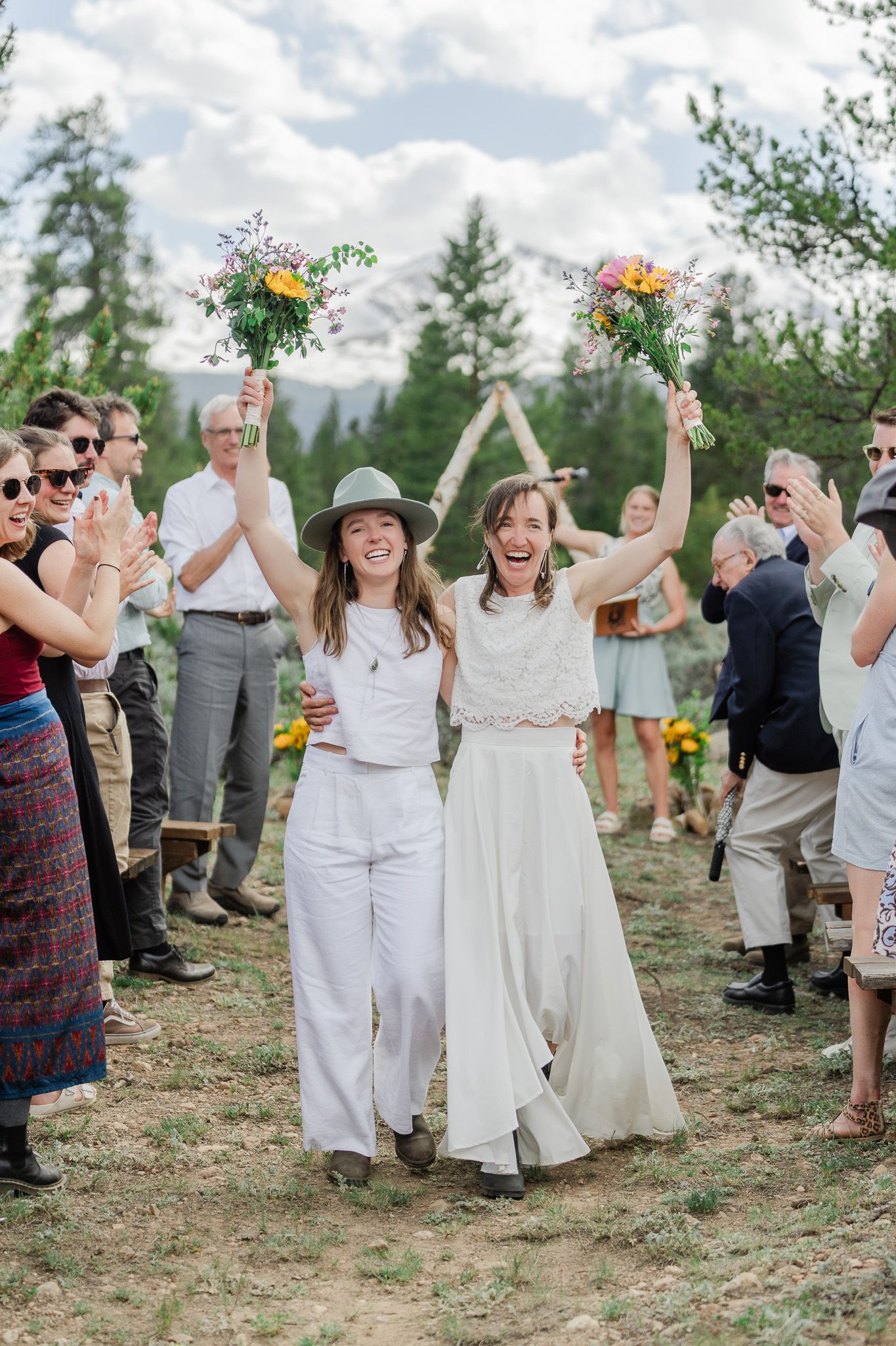 Two brides in white celebrating joyfully with raised bouquets after outdoor mountain wedding ceremony.