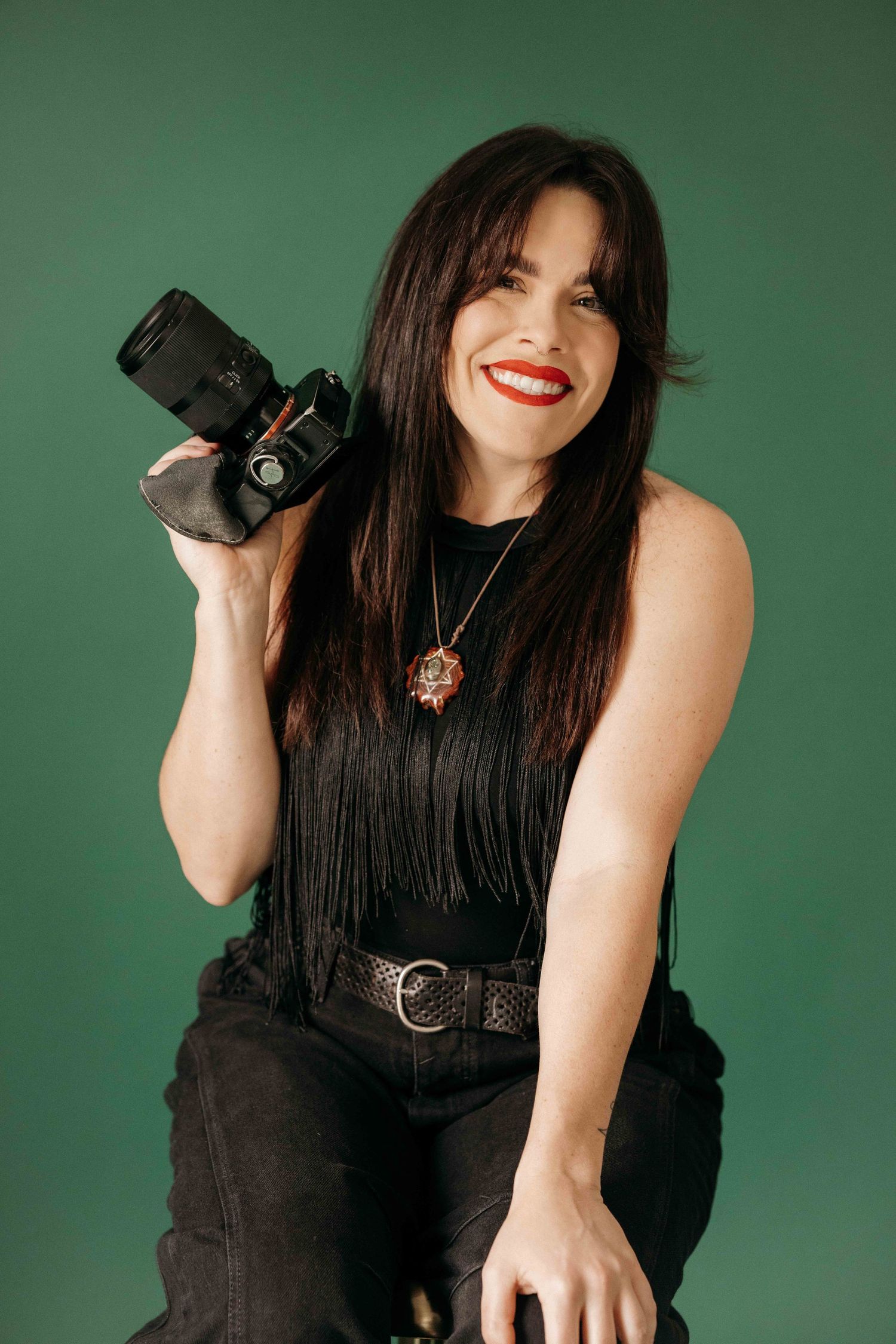 Smiling brunette woman in black fringe top holding a DSLR camera against a green studio background.