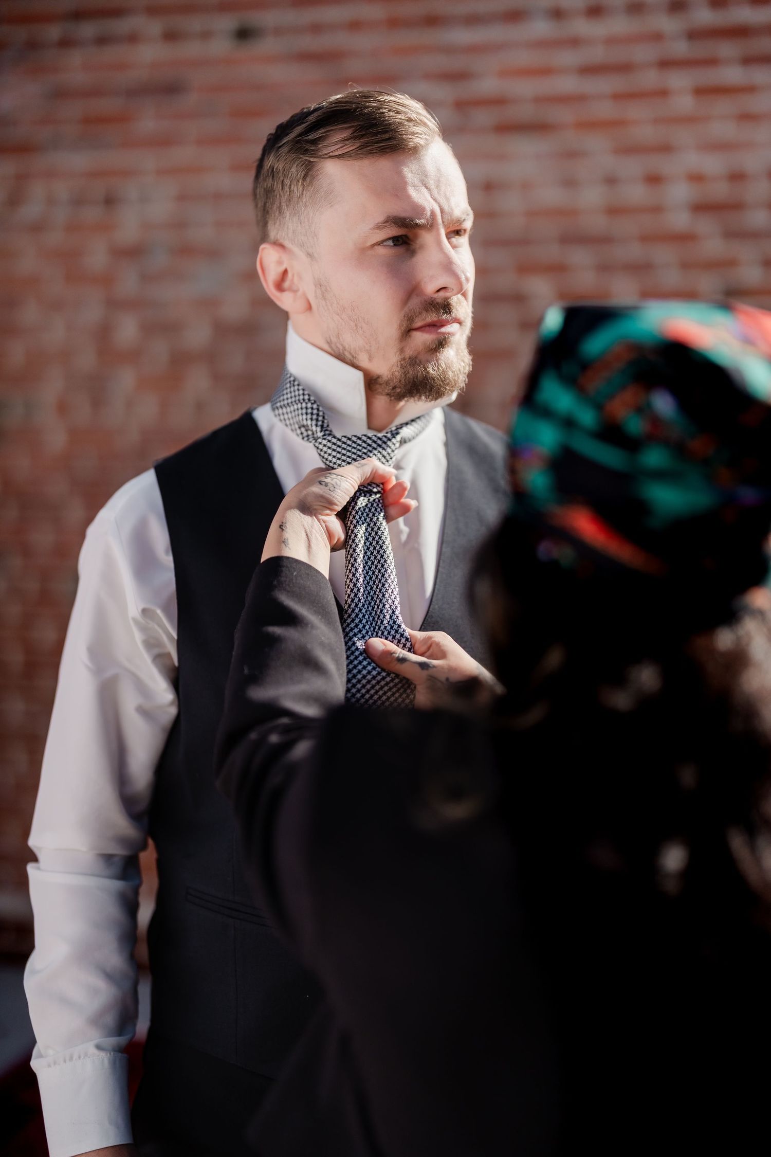 A woman adjusts a man's tie as he dresses in a white shirt and dark vest near a brick wall.
