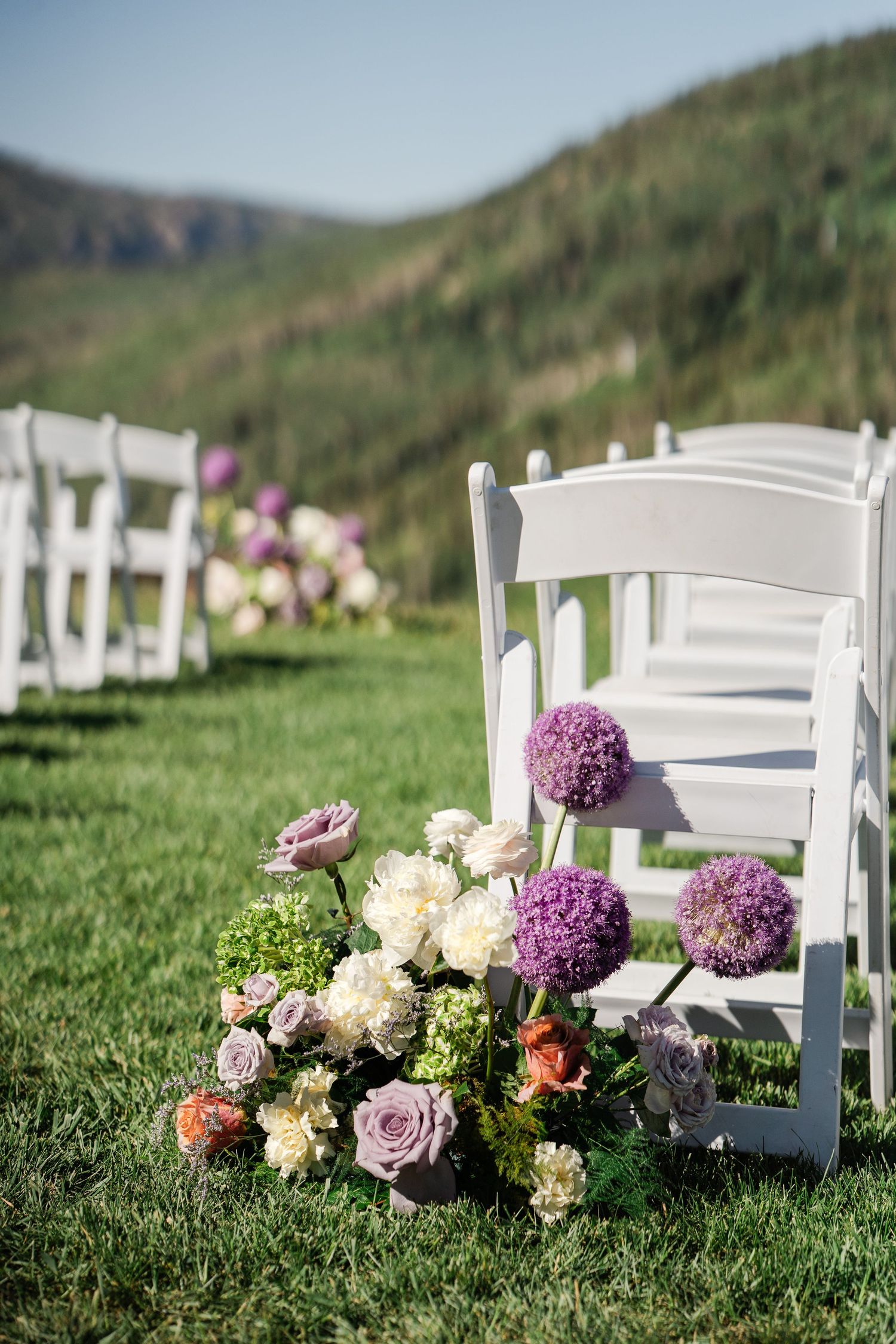 Purple allium and rose floral arrangements lining white chairs at an outdoor mountain wedding.