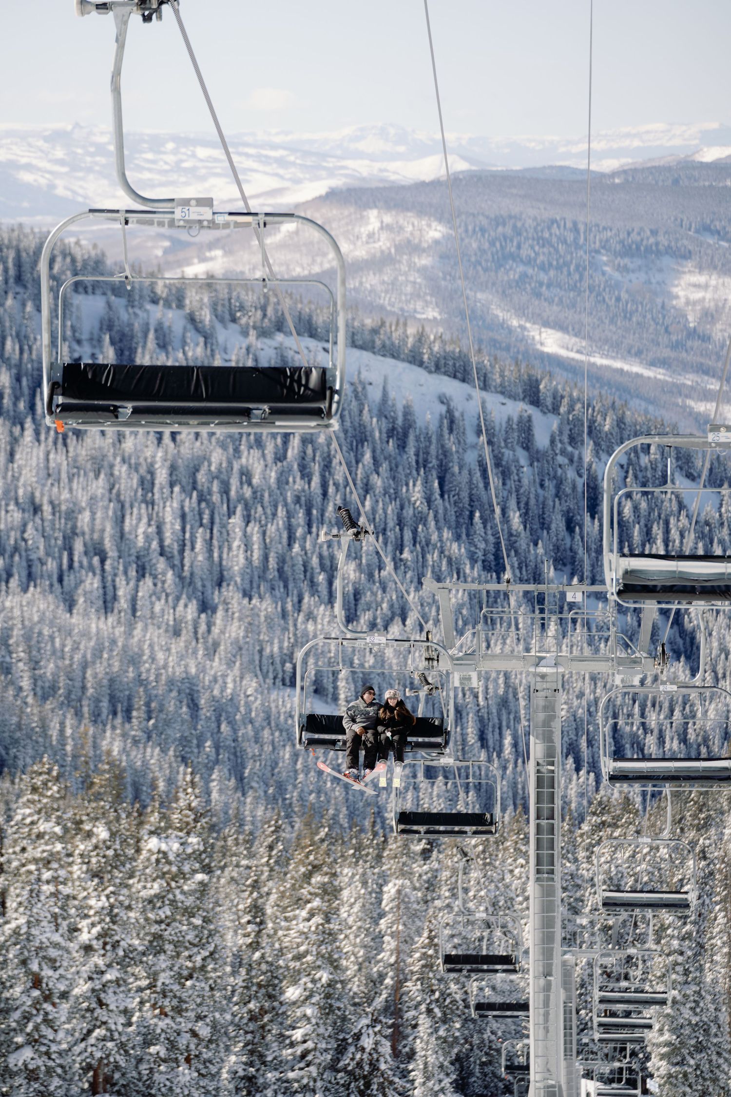 Ski lift chairs ascending snowy mountain slope with frost-covered pine trees and mountain range backdrop.