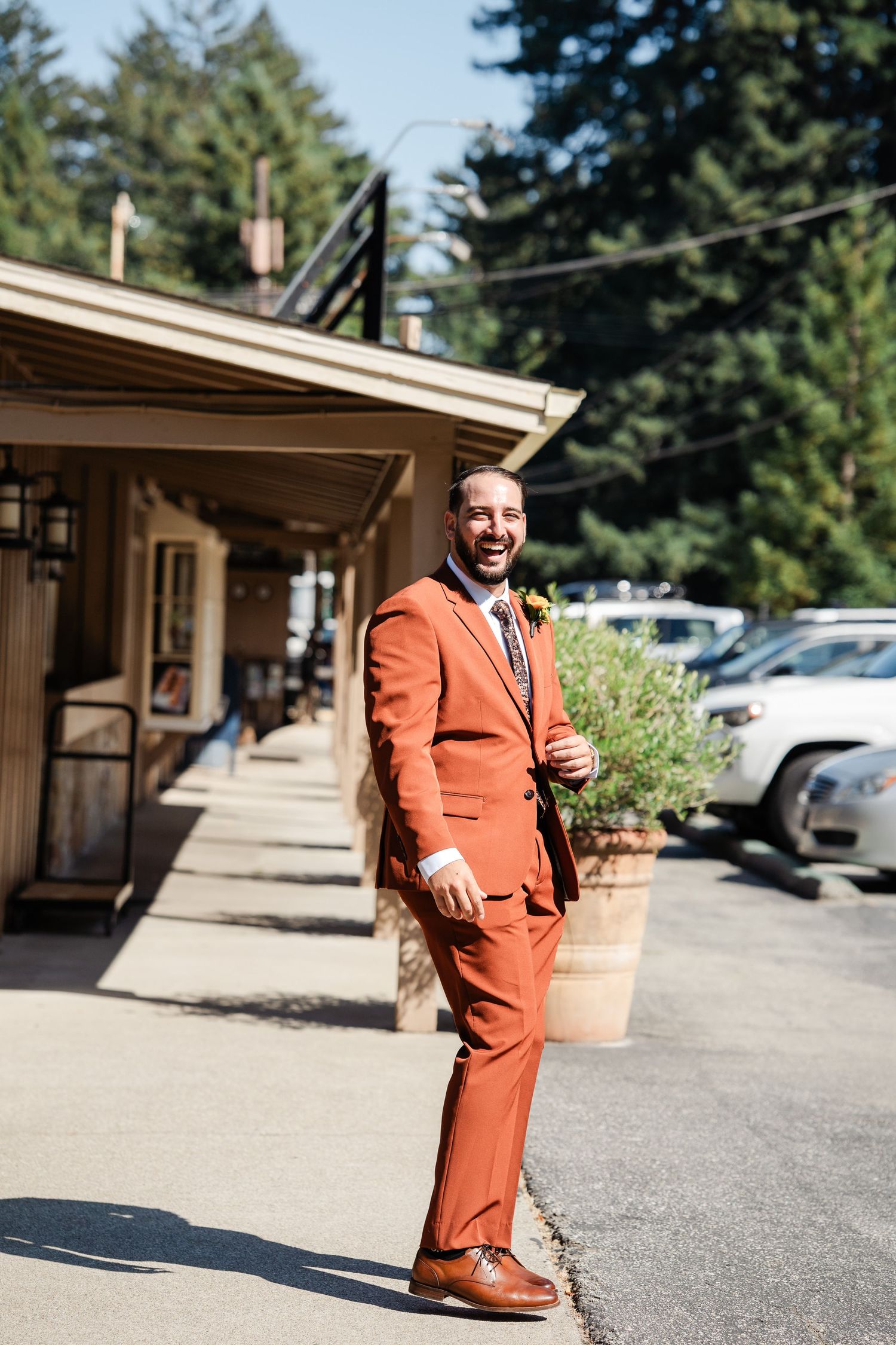 Smiling groom in rust orange suit with boutonniere posing outside rustic venue building.
