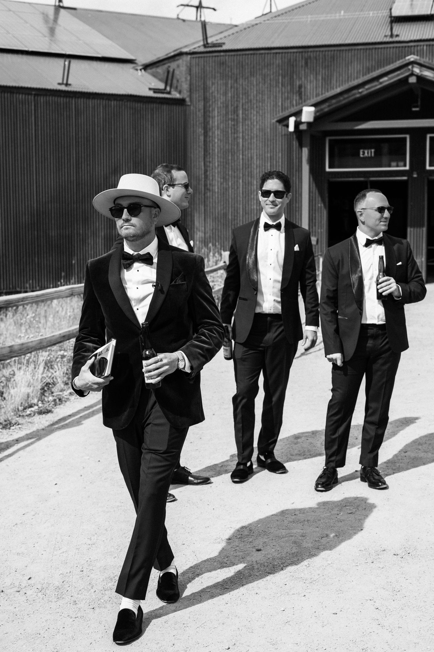 Stylish groomsmen in black tuxedos and one wearing white hat walking outdoors in black and white photo.