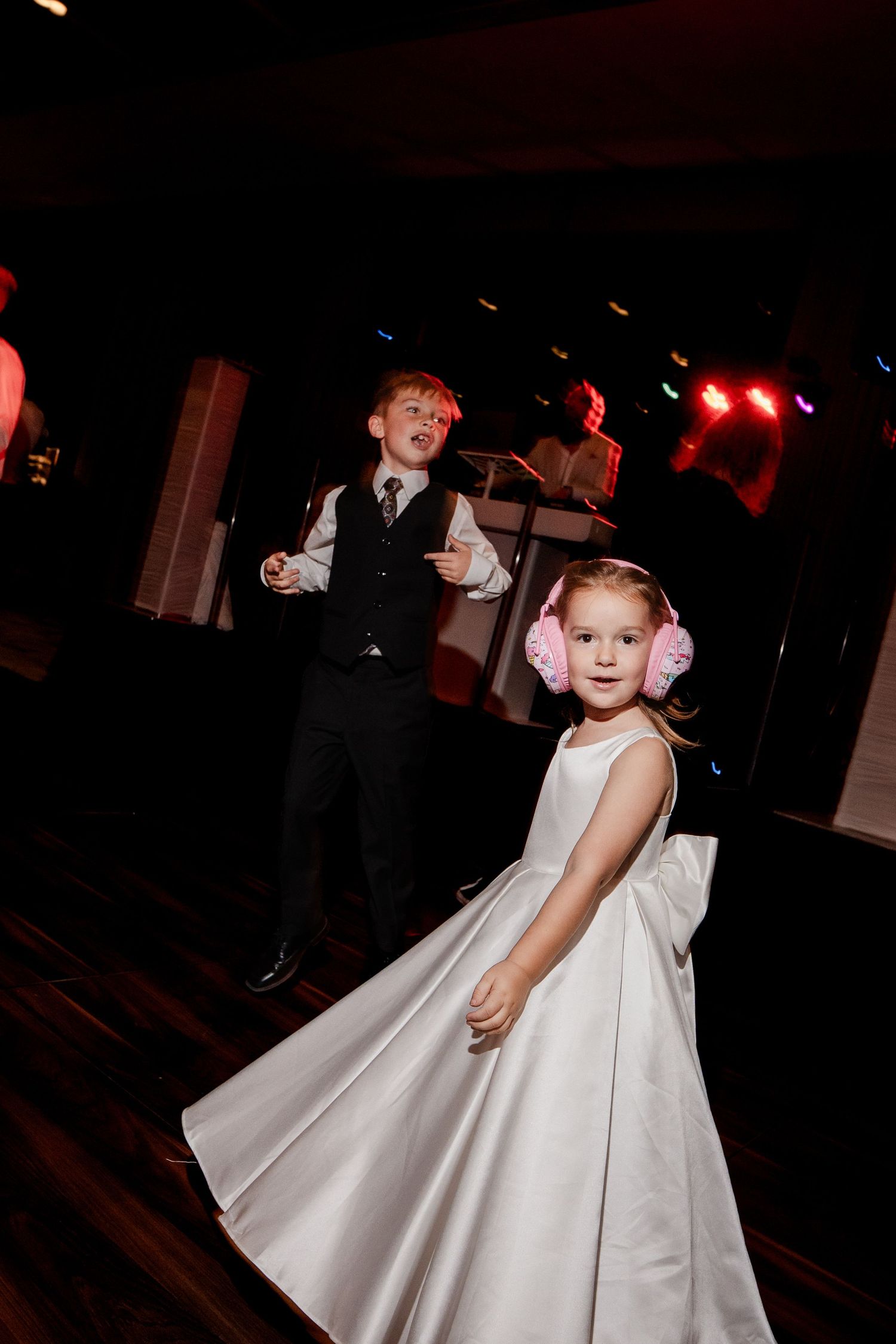 Young ring bearer and flower girl with pink earmuffs dancing joyfully at wedding reception.