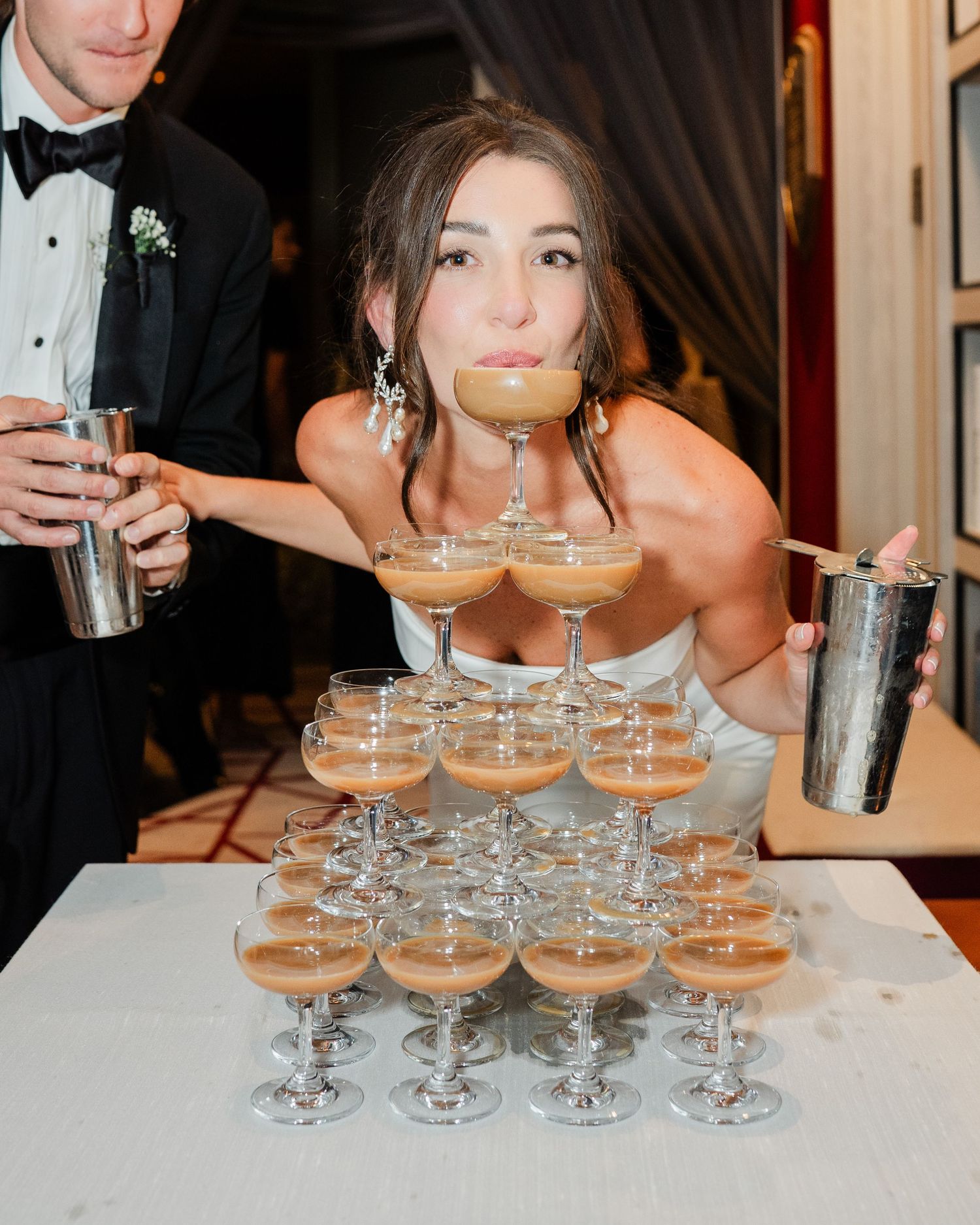 Bride leans over a tiered espresso martini coupe glass tower at wedding reception.