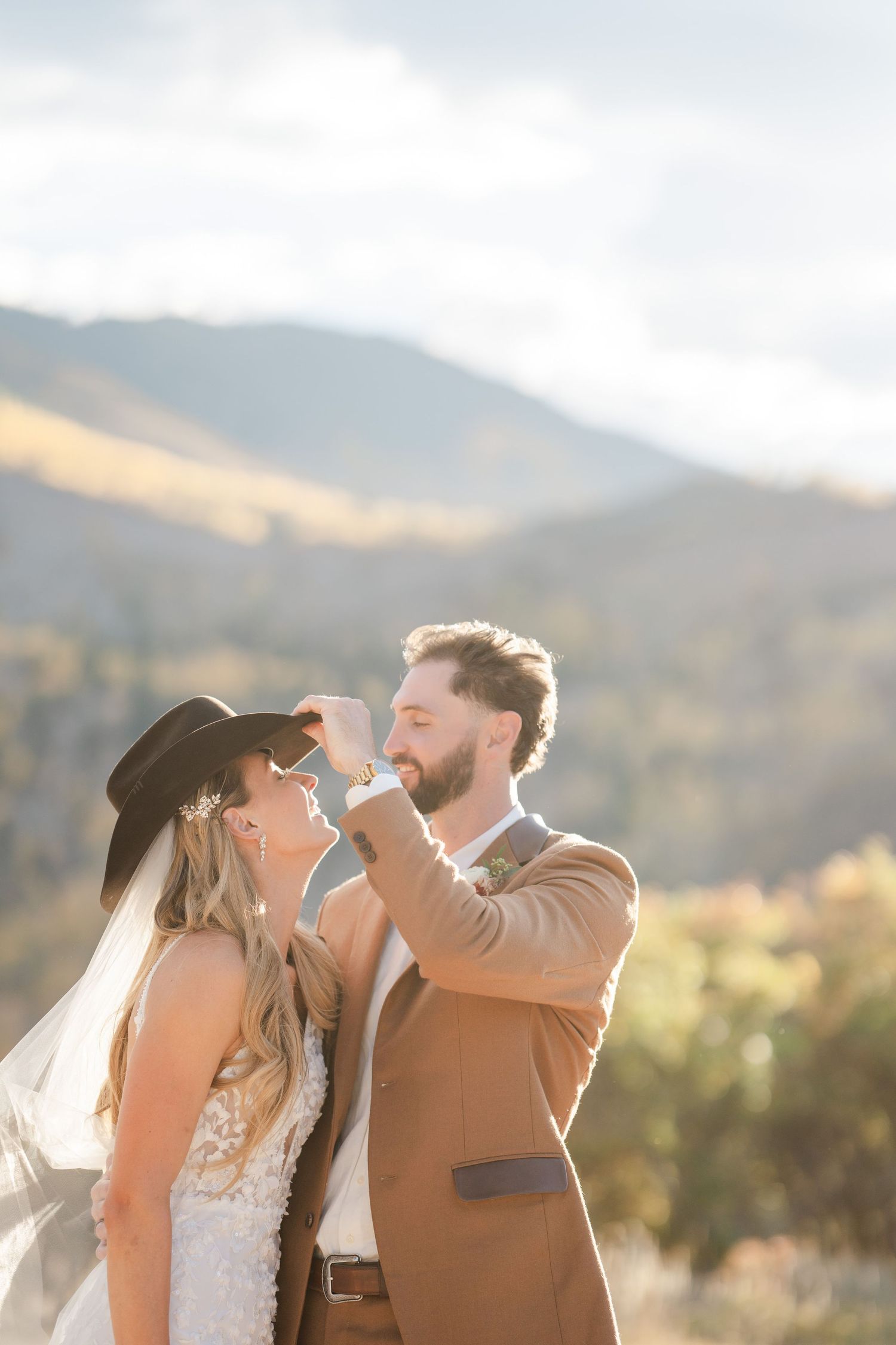 A groom in a tan suit playfully tips his bride's cowboy hat during a sunny mountain wedding.