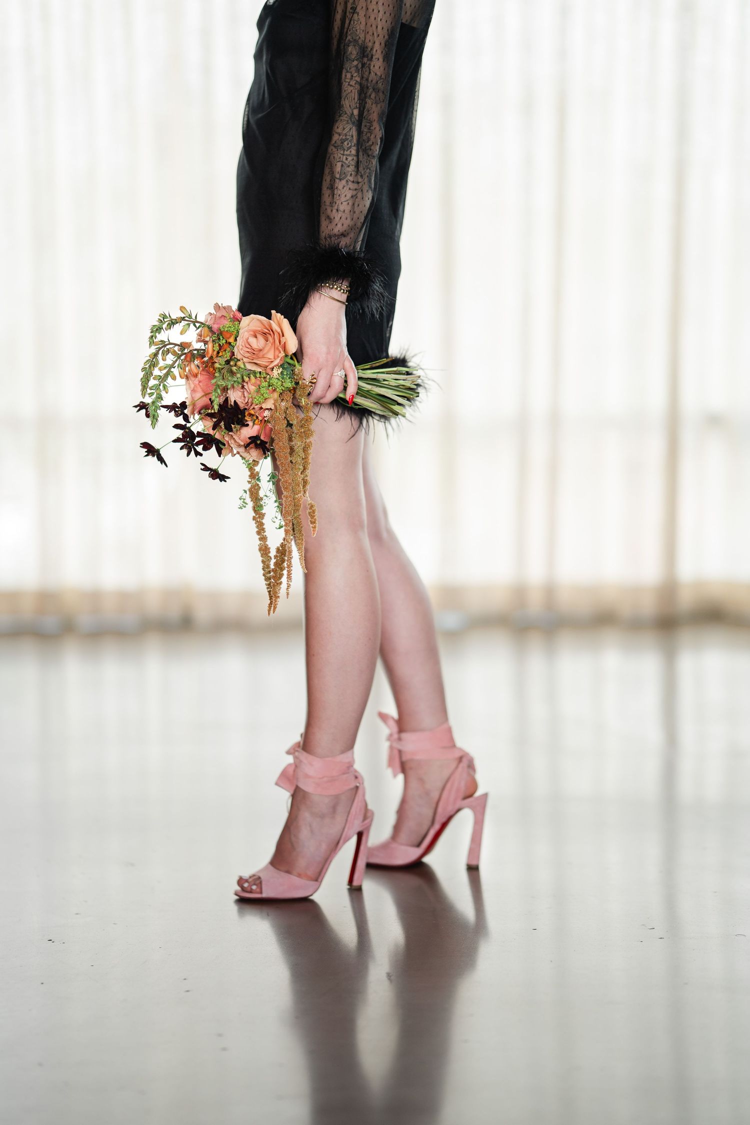 A bride in a black lace dress holds a coral bouquet while wearing blush pink heeled sandals indoors.