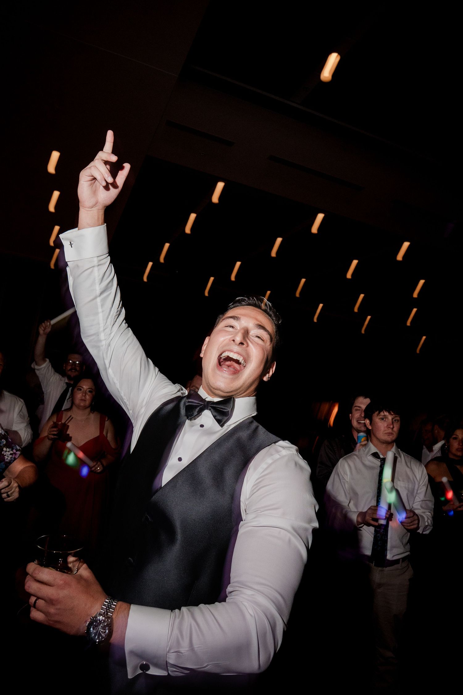 Excited groomsman in tuxedo vest celebrating with arm raised on crowded wedding reception dance floor.