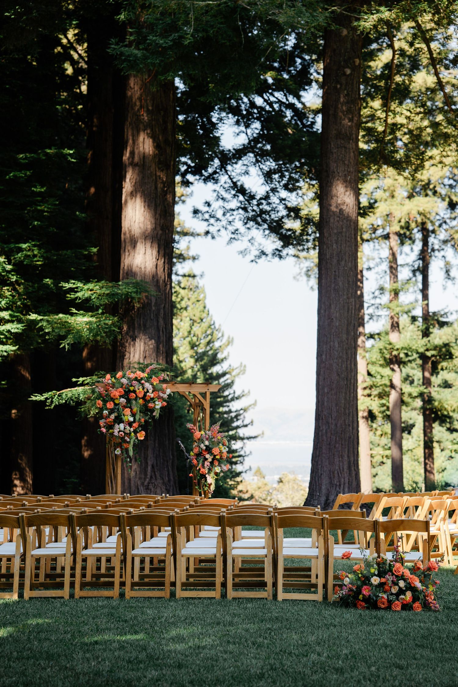 Elegant outdoor wedding ceremony with wooden arch, orange floral arrangements, and redwood trees.