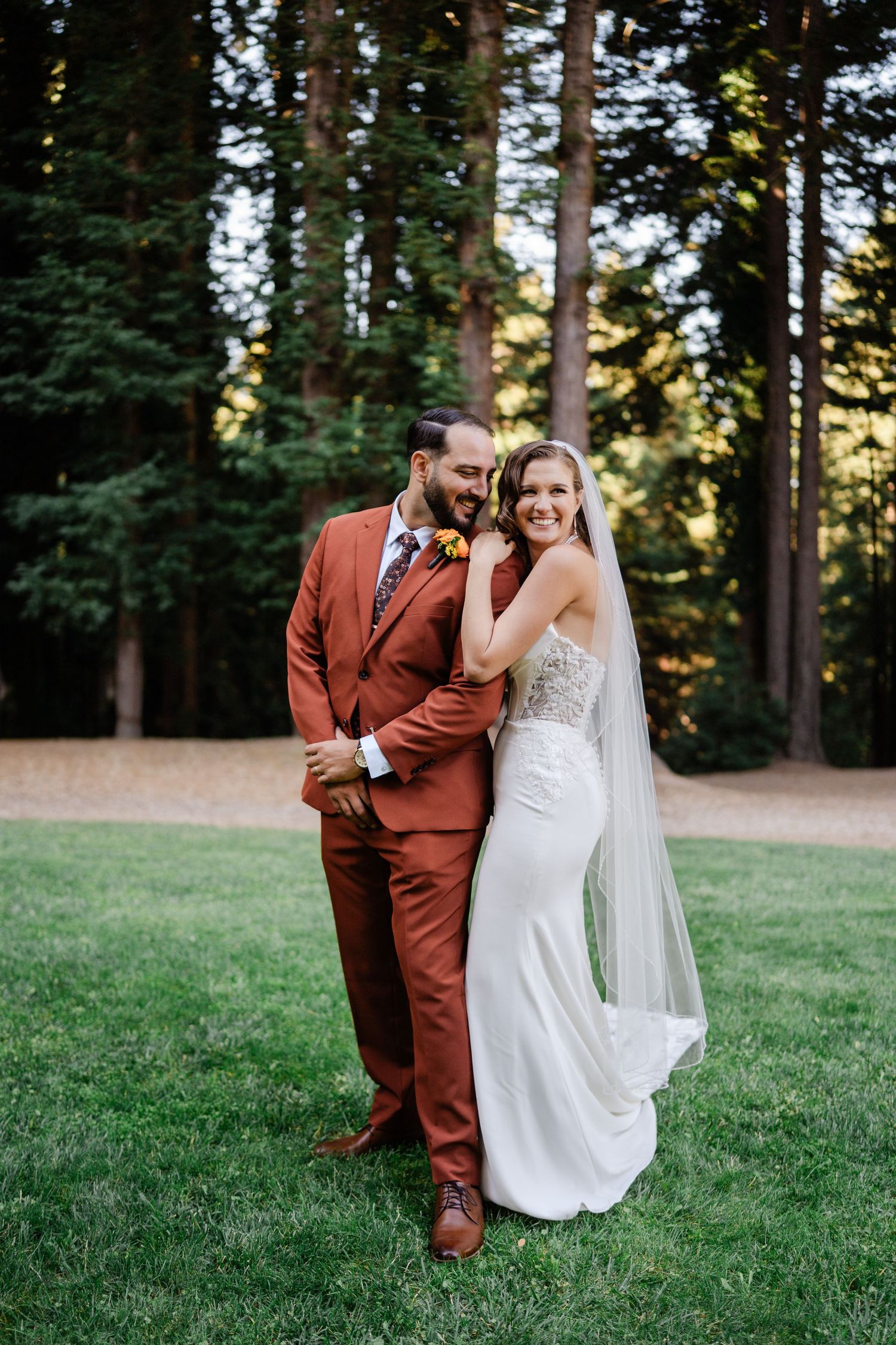 Bride and groom posing in a lush forest setting, groom in rust suit, bride in fitted white gown.