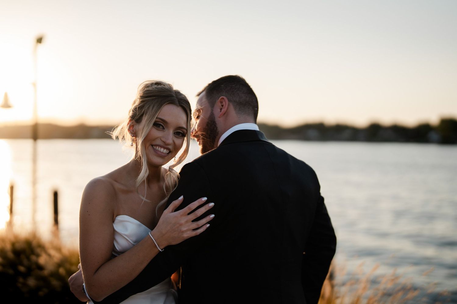 Bride smiling at groom during golden hour sunset portrait by a serene waterfront location.