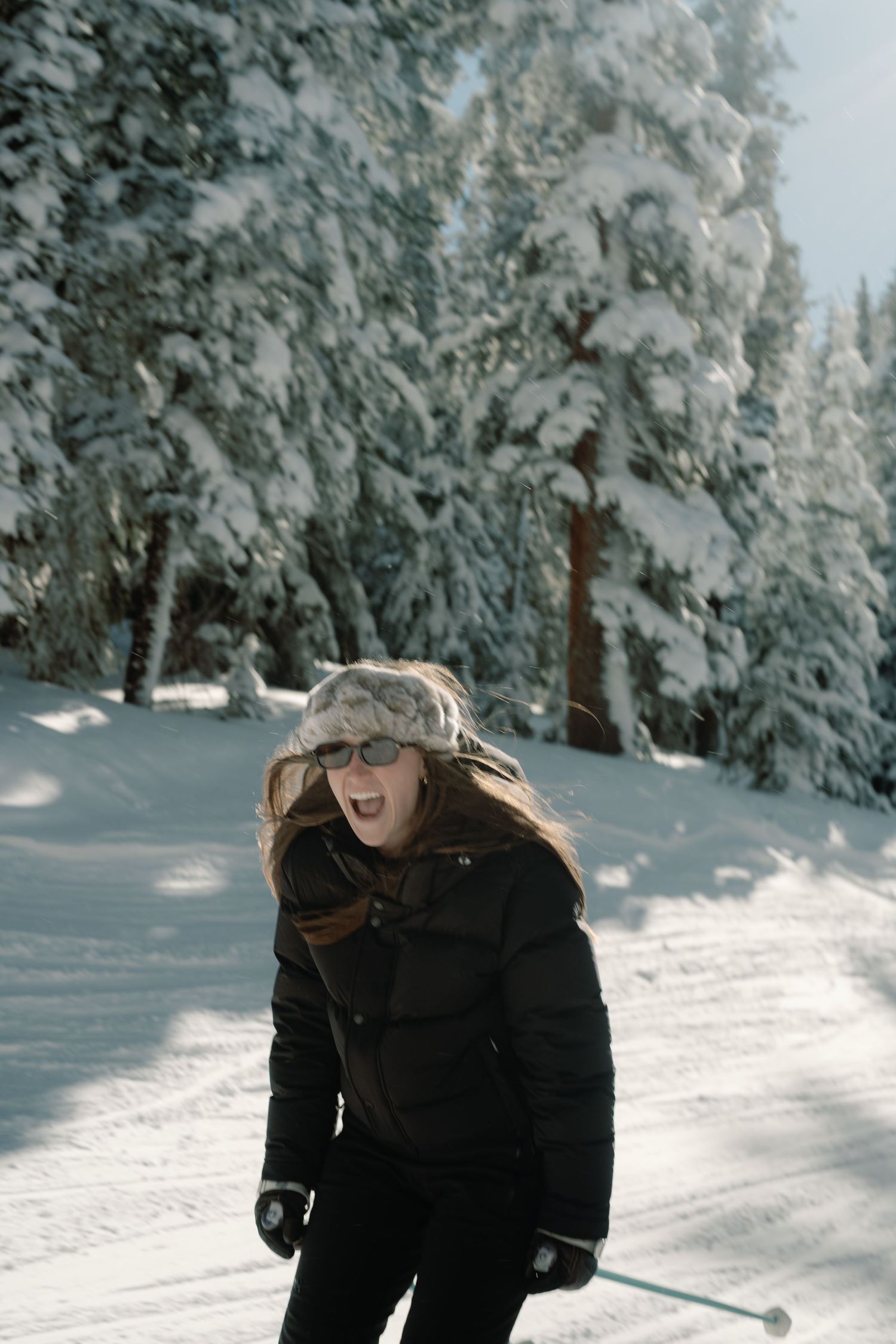 Woman in fur hat and sunglasses skiing through snow-covered pine forest on sunny day.