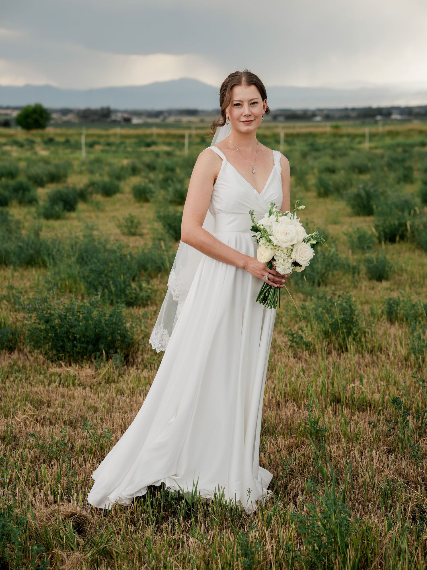 Bride in flowing white V-neck gown holding white bouquet in an open field with mountain views.