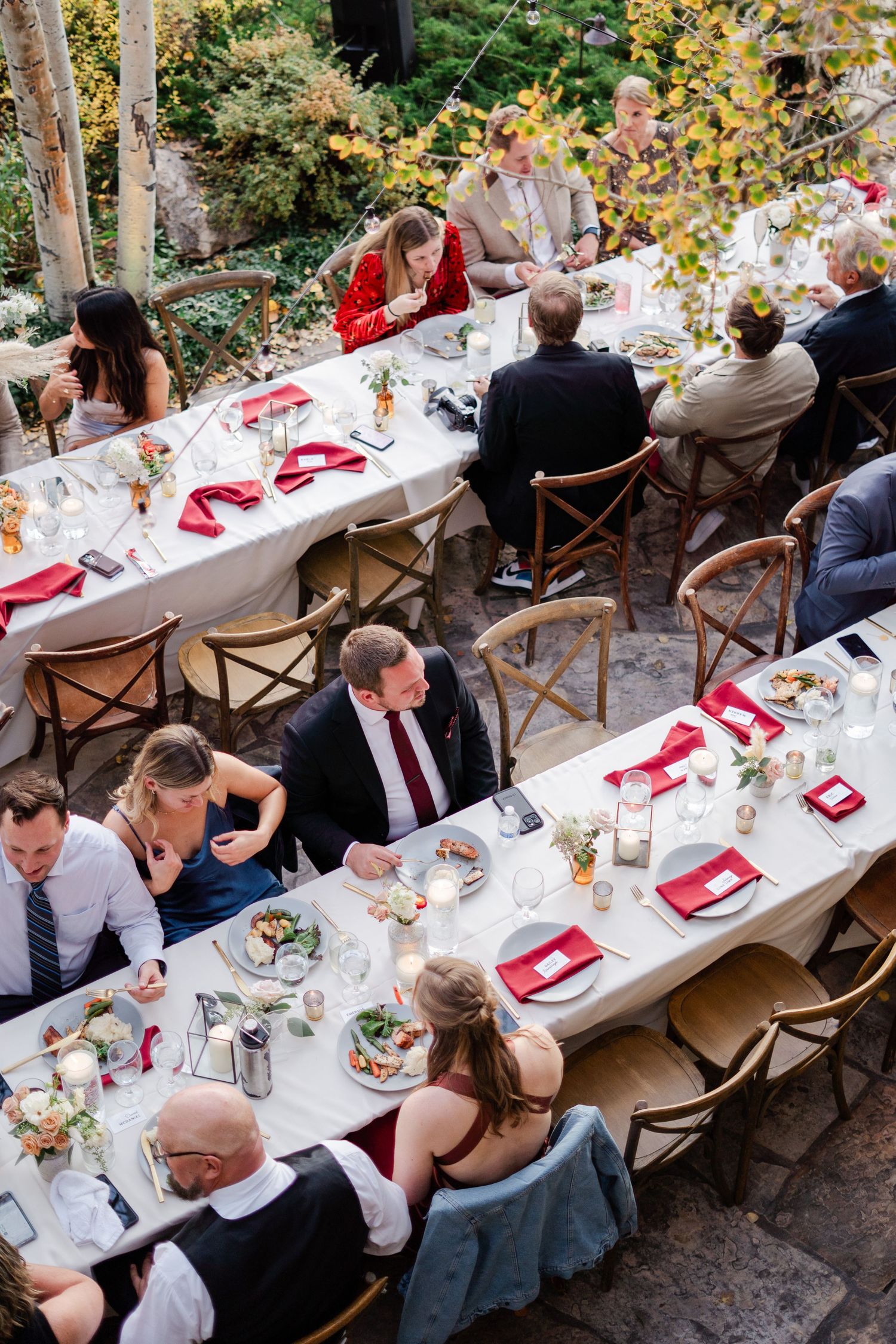 Aerial view of wedding guests dining at long tables with red napkins at an outdoor fall reception.