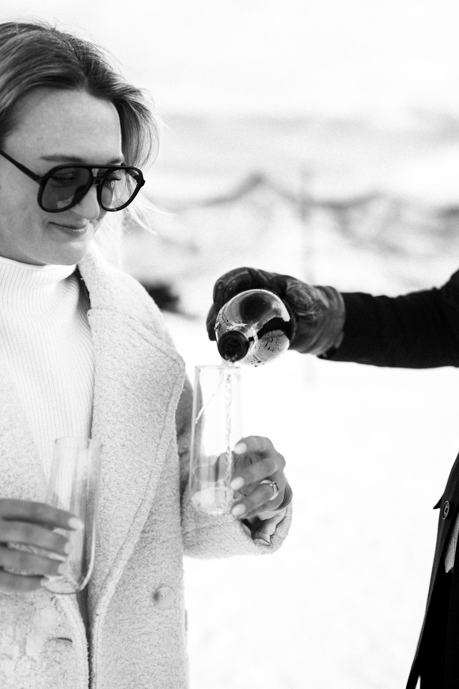 Black and white photo of woman in white coat receiving champagne pour on snowy mountain.