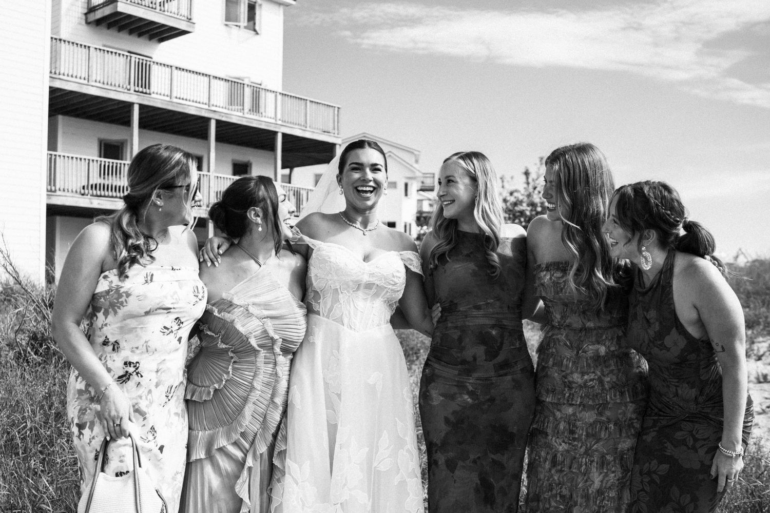 Black and white photo of smiling bride surrounded by five bridesmaids outdoors at coastal wedding venue.