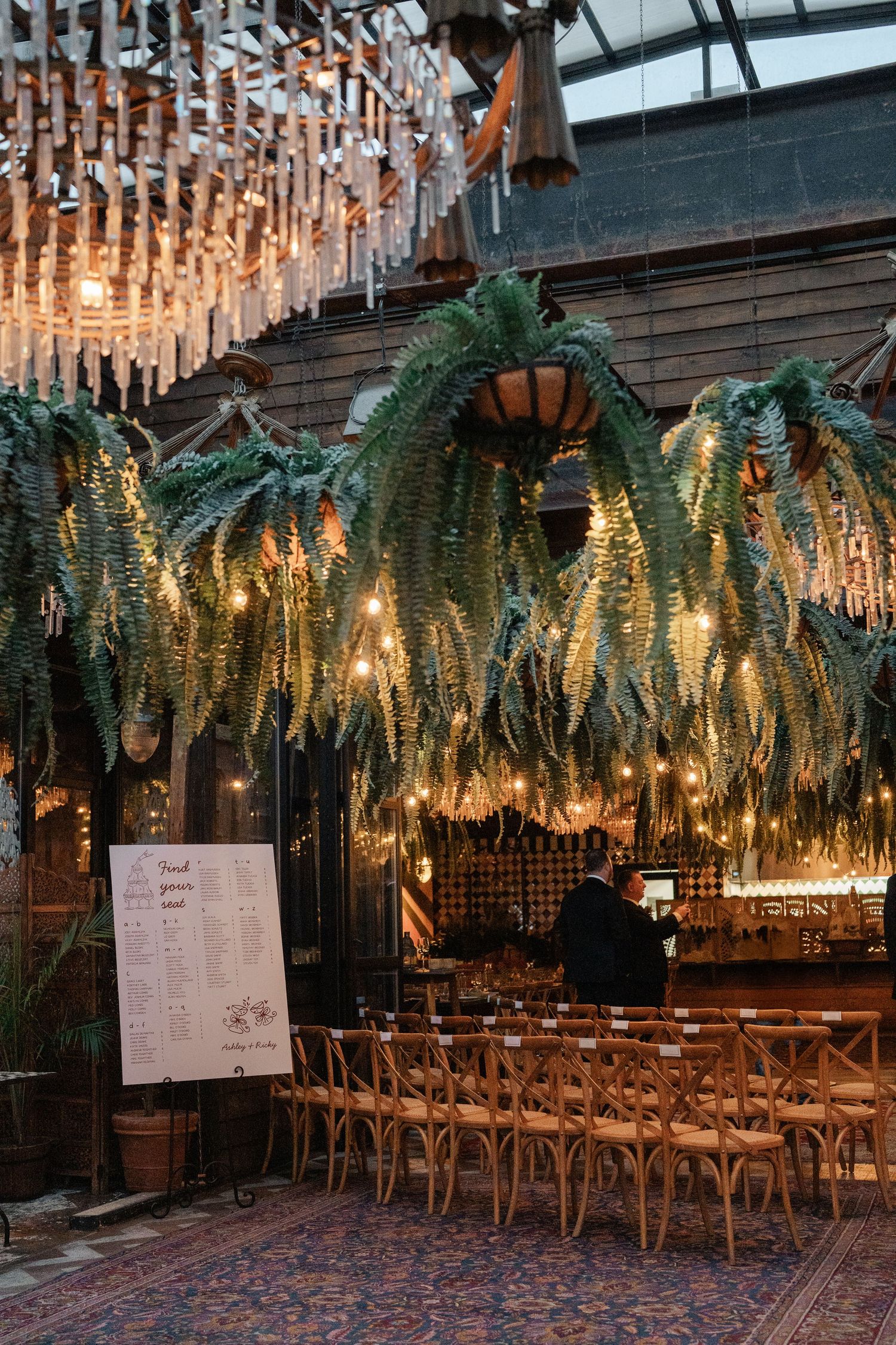 Rustic wedding venue interior with hanging ferns, string lights, and wooden cross-back chairs.