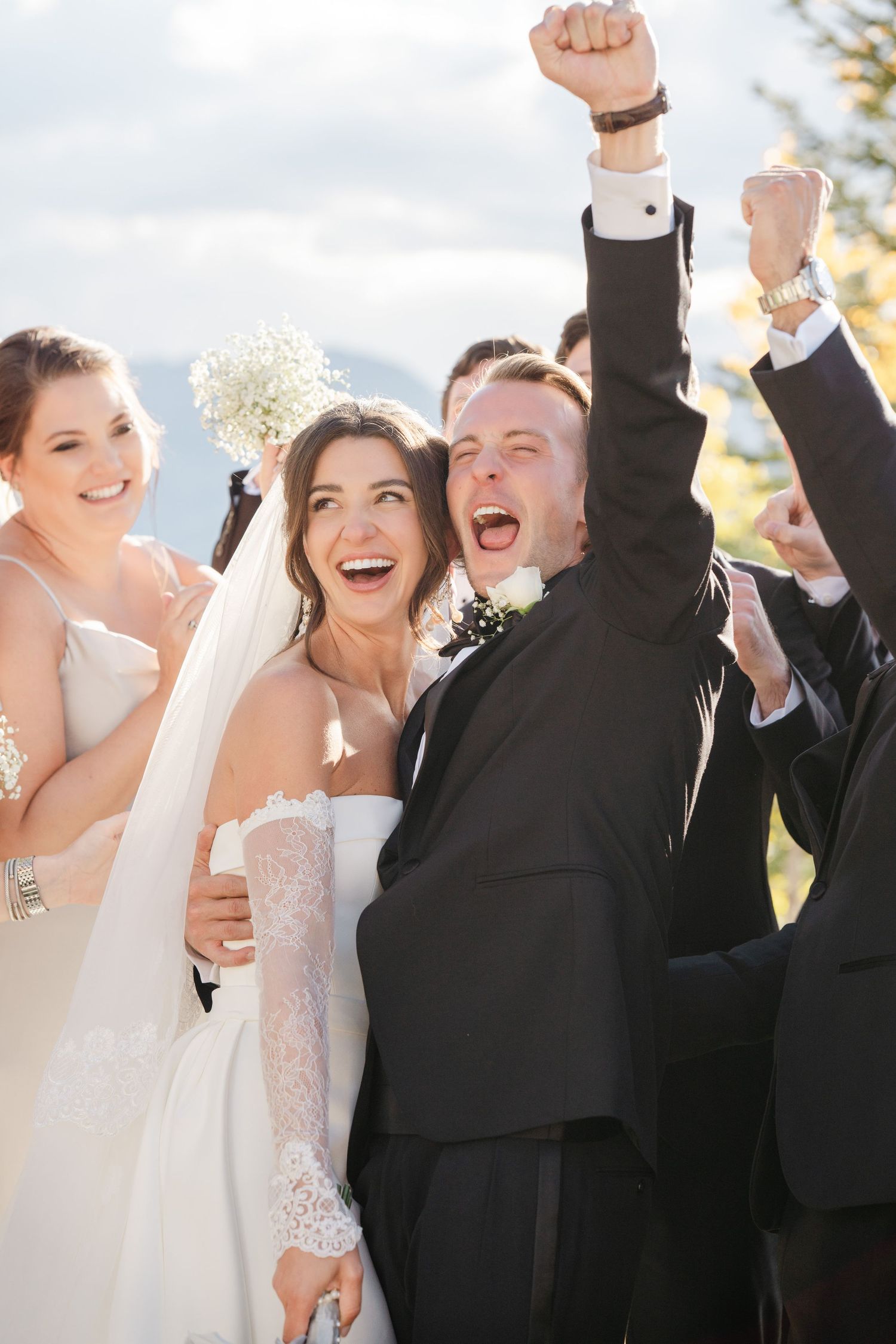 Elated bride and groom celebrate with wedding party as groom raises fist in joy at outdoor mountain wedding.