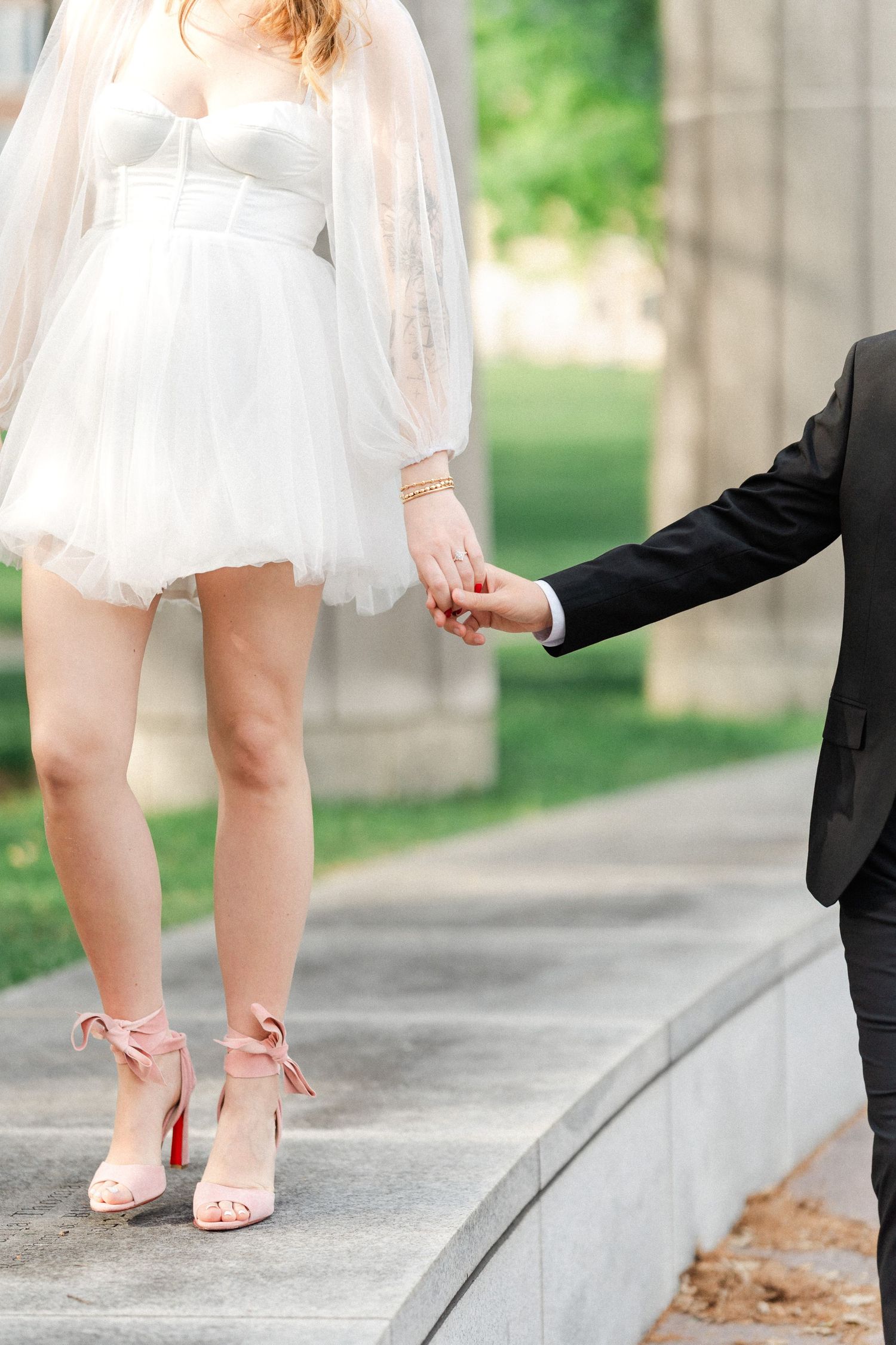 Bride in short white tulle dress with pink bow heels holding grooms hand on stone steps outdoors.