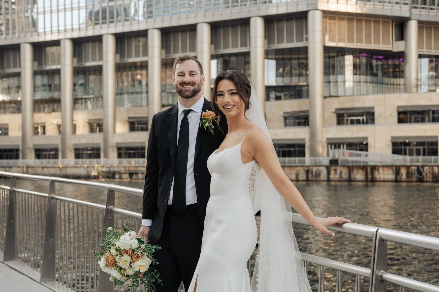 Smiling newlyweds posing on urban waterfront bridge with bouquet and city building backdrop.