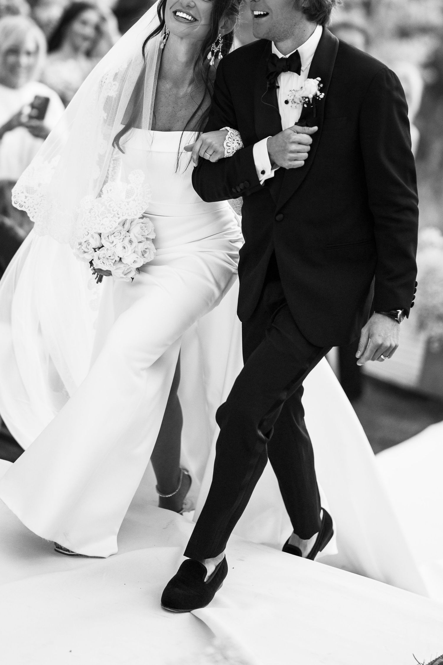 Bride and groom walk together joyfully after wedding ceremony, bride holding white bouquet in elegant white gown.