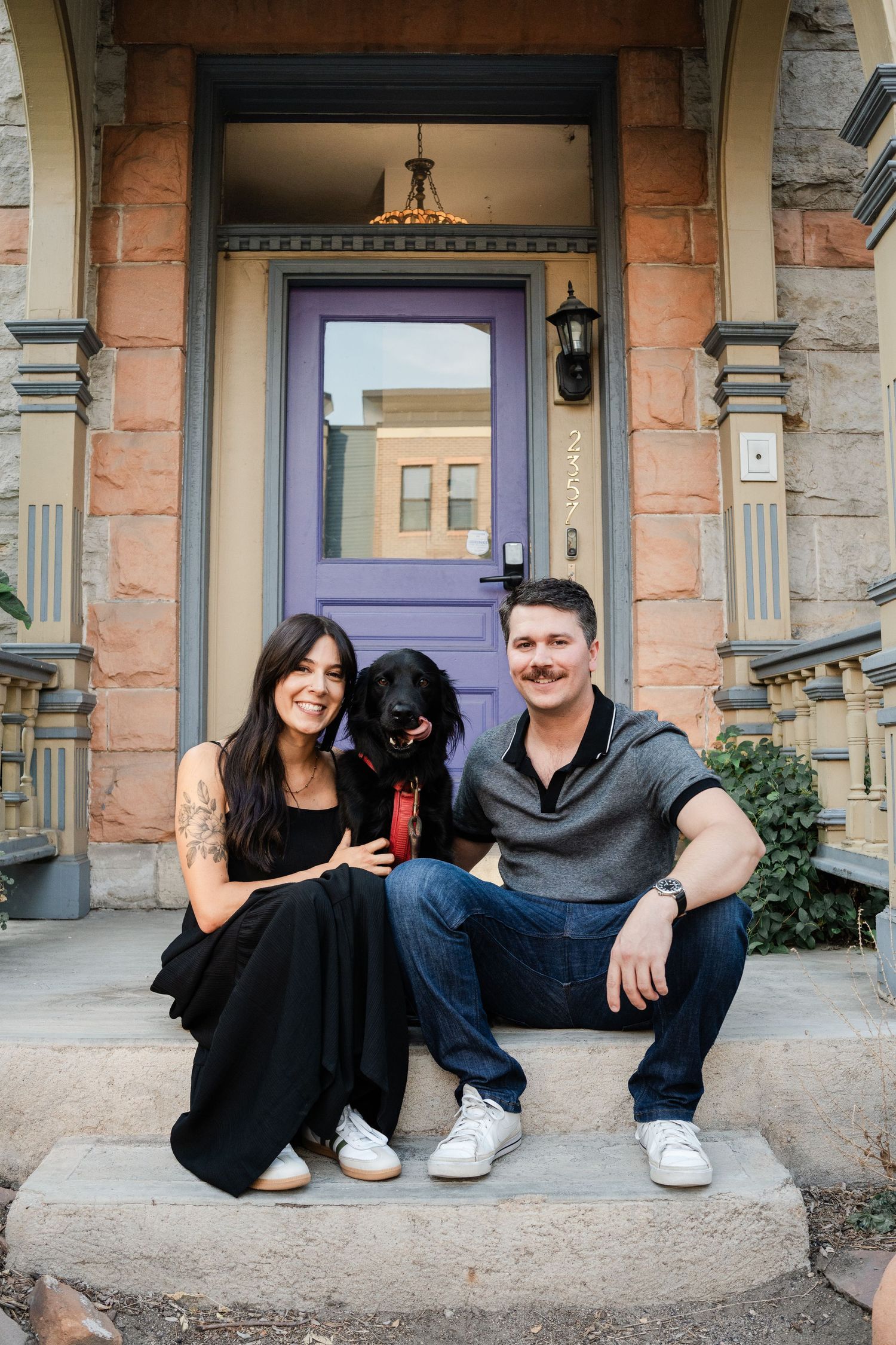 Smiling couple with black dog sitting on steps of home with purple front door.