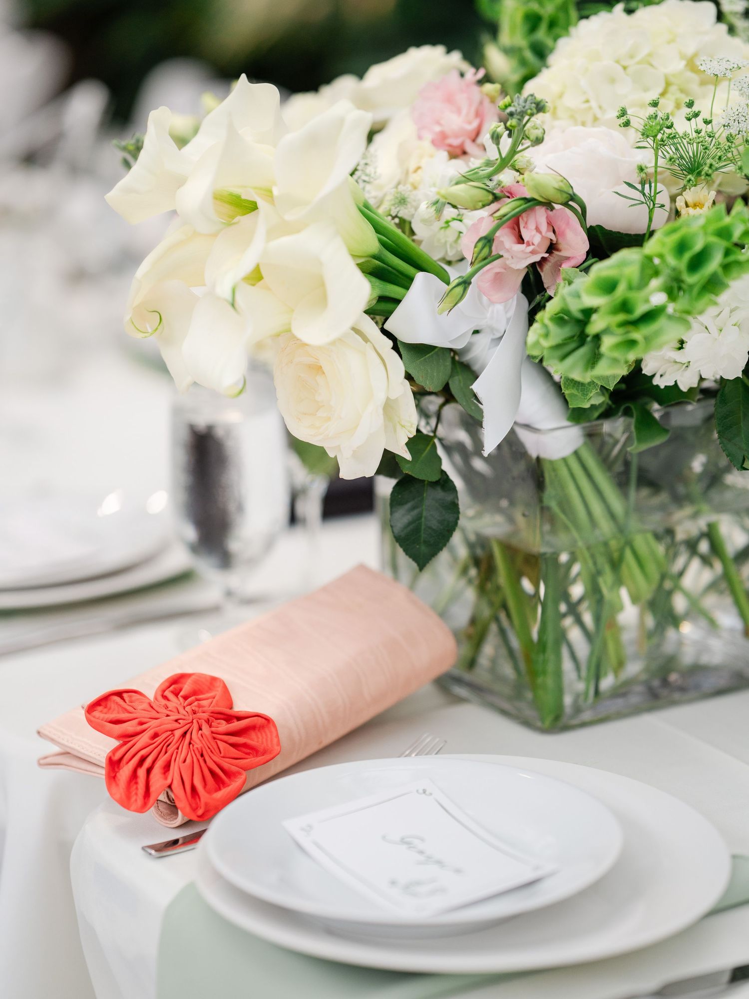 Elegant wedding table setting with white and pink floral centerpiece, coral napkin accent, and white place settings.