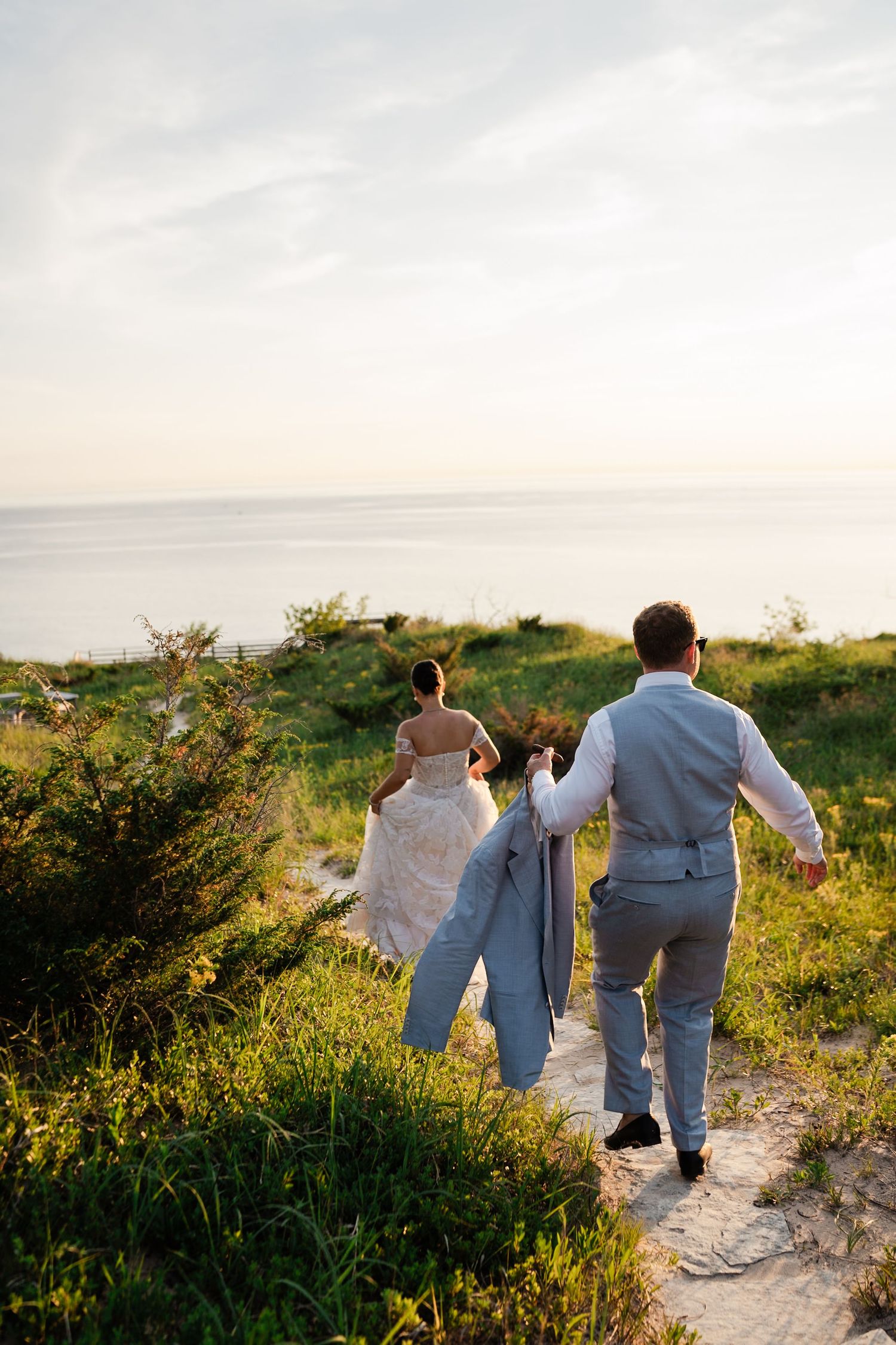 Bride and groom walking hand in hand toward the ocean on a grassy coastal path at sunset.